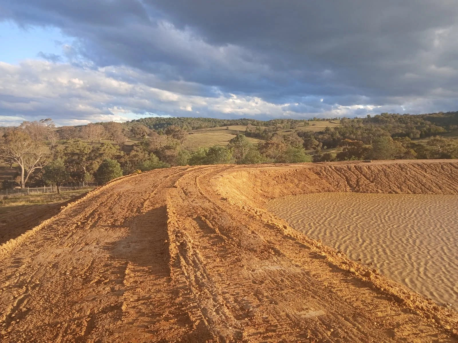 A dirt road curves through a sandy, open landscape with a backdrop of rolling hills and scattered trees under a partly cloudy sky during daylight.