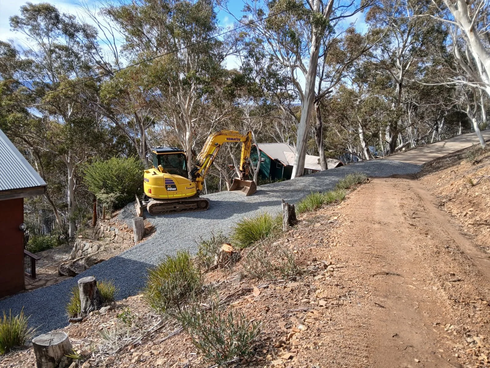A small yellow excavator working on a slope covered with gravel, surrounded by trees closest to a dirt path in Snowy Mountains NSW