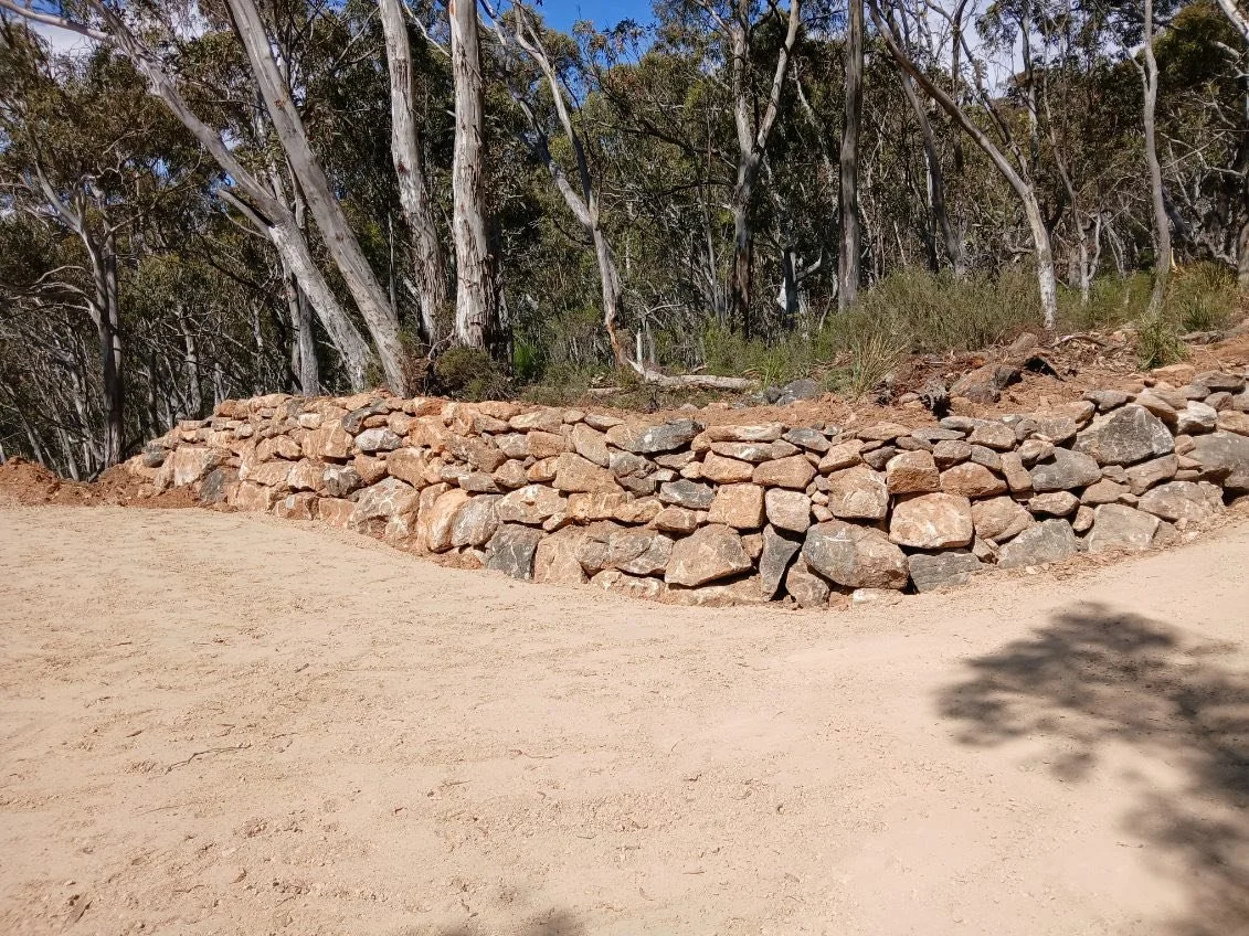A low stone wall made of irregularly shaped rocks in front of a dirt path with a forest of tall, thin trees in the background. Landscaping done in Jindabyne 2627