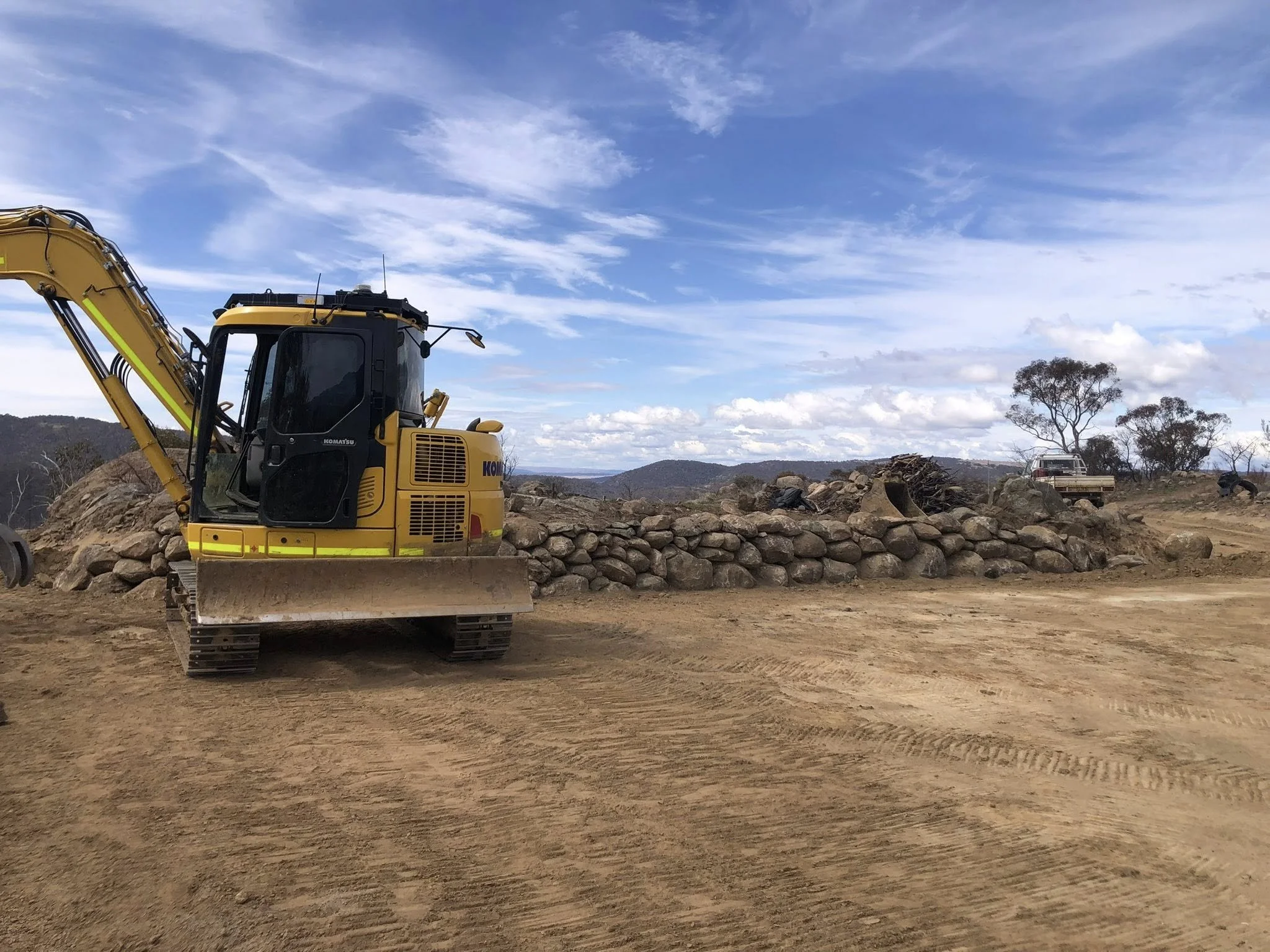 Construction site with a yellow excavator and a rock wall under a partly cloudy sky.
