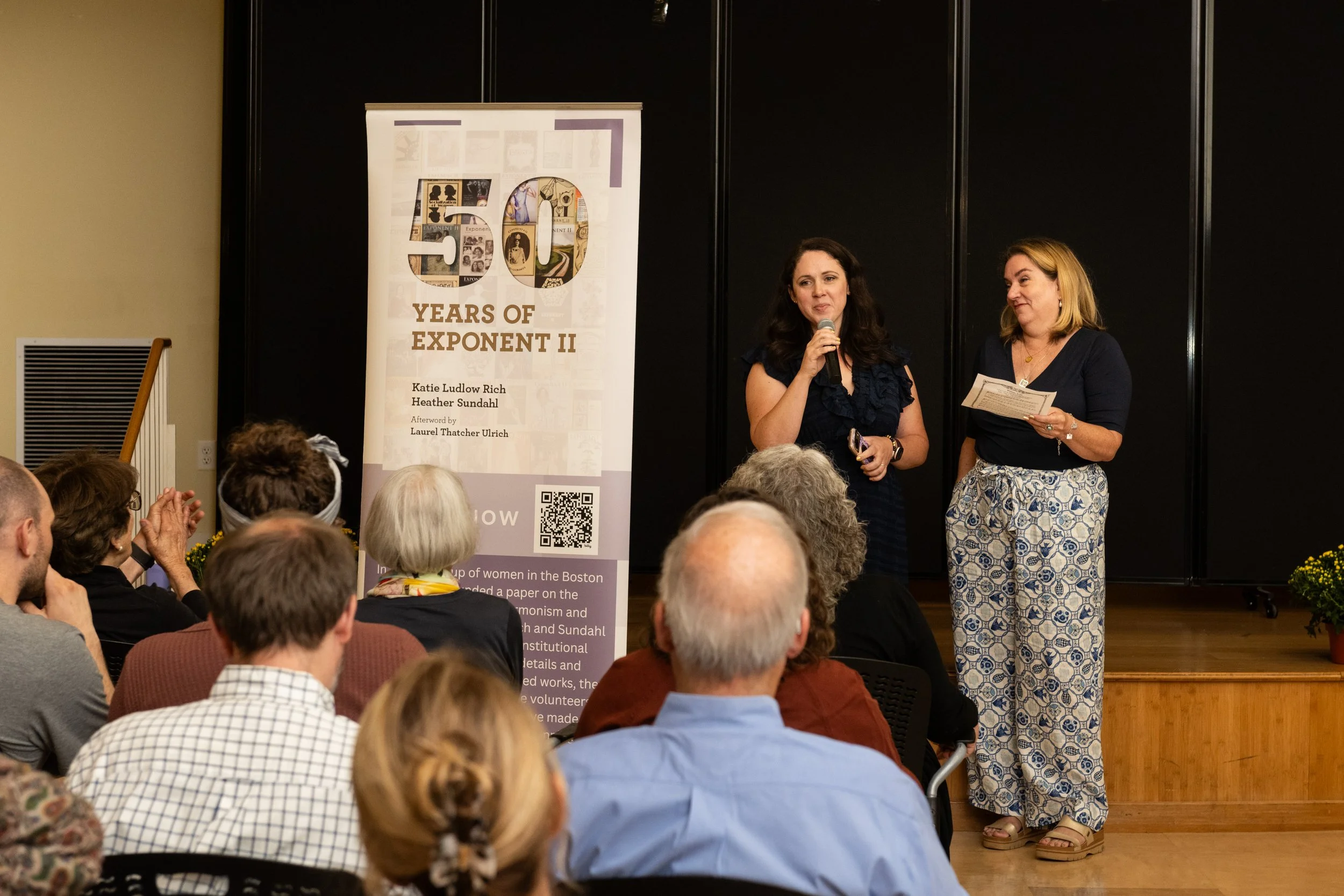 Katie Ludlow Rich and Heather Sundahl standing on stage next to a vertical banner at an Exponent II event. The audience is seated facing the stage, and the banner reads :50 Years of Exponent II" with additional text below.