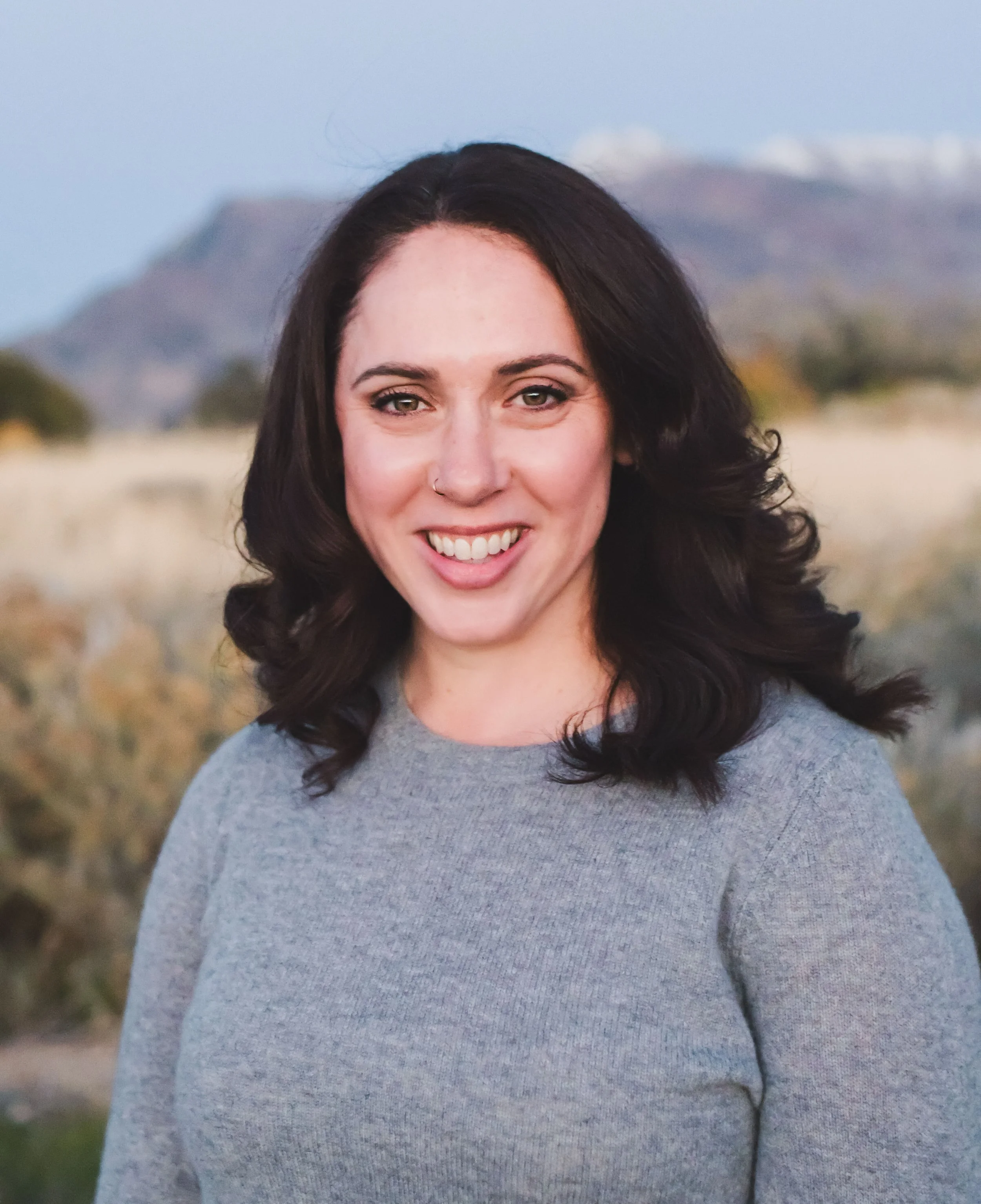 Historian Katie Ludlow Rich is pictured with dark hair, smiling, wearing a gray top, outdoors with a mountainous background.
