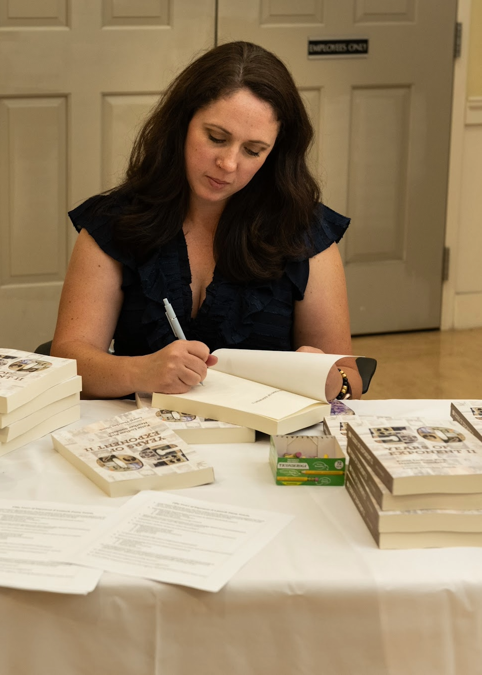 Katie Ludlow Rich sitting at a table signing her book, Fifty Years of Exponent II. Several stacks of books and a few sheets of paper are on the table.