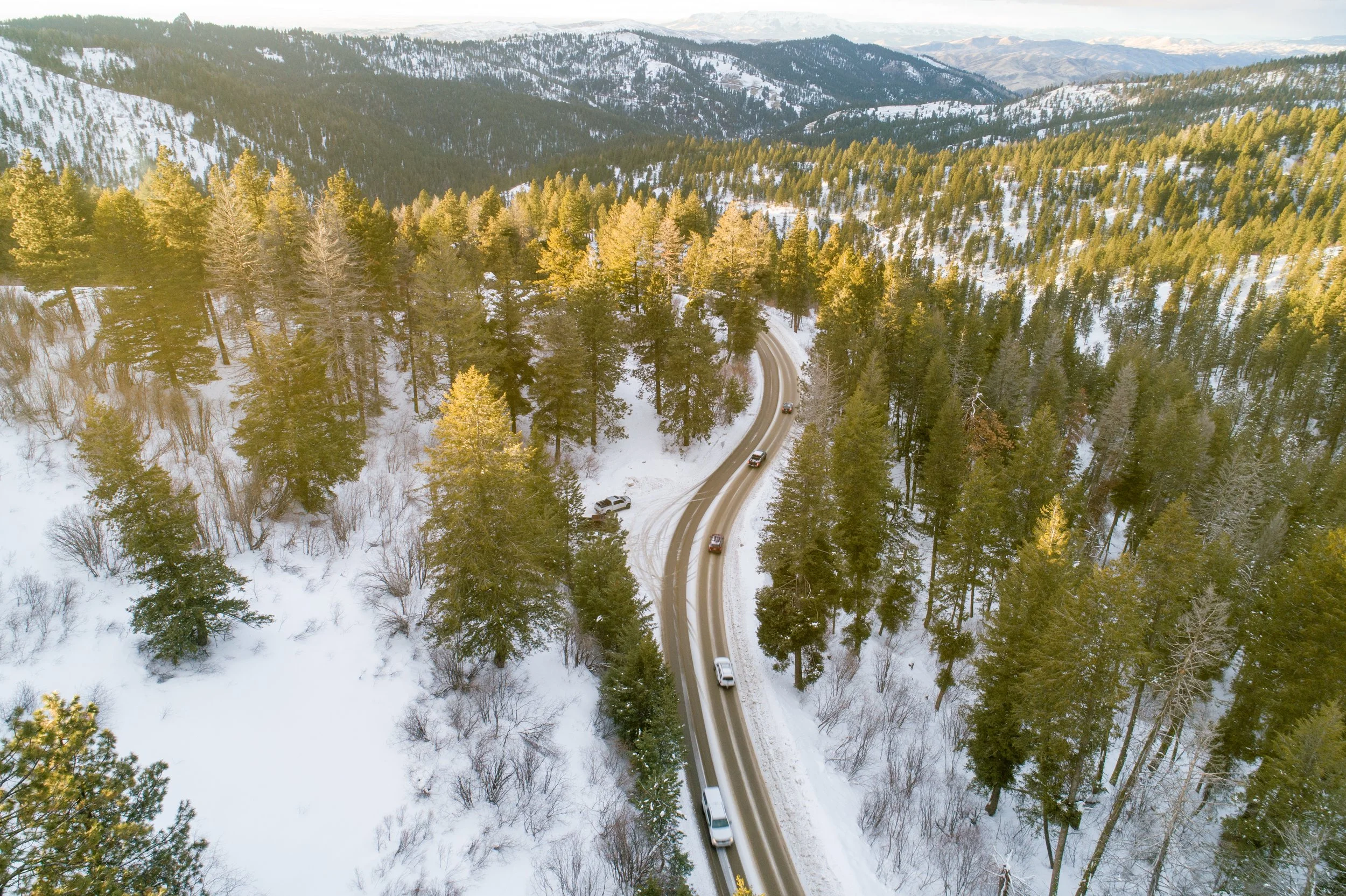 Aerial view of a winding snowy mountain road through a dense forest of evergreen trees near McCall, with snow-covered mountain peaks in the distance.