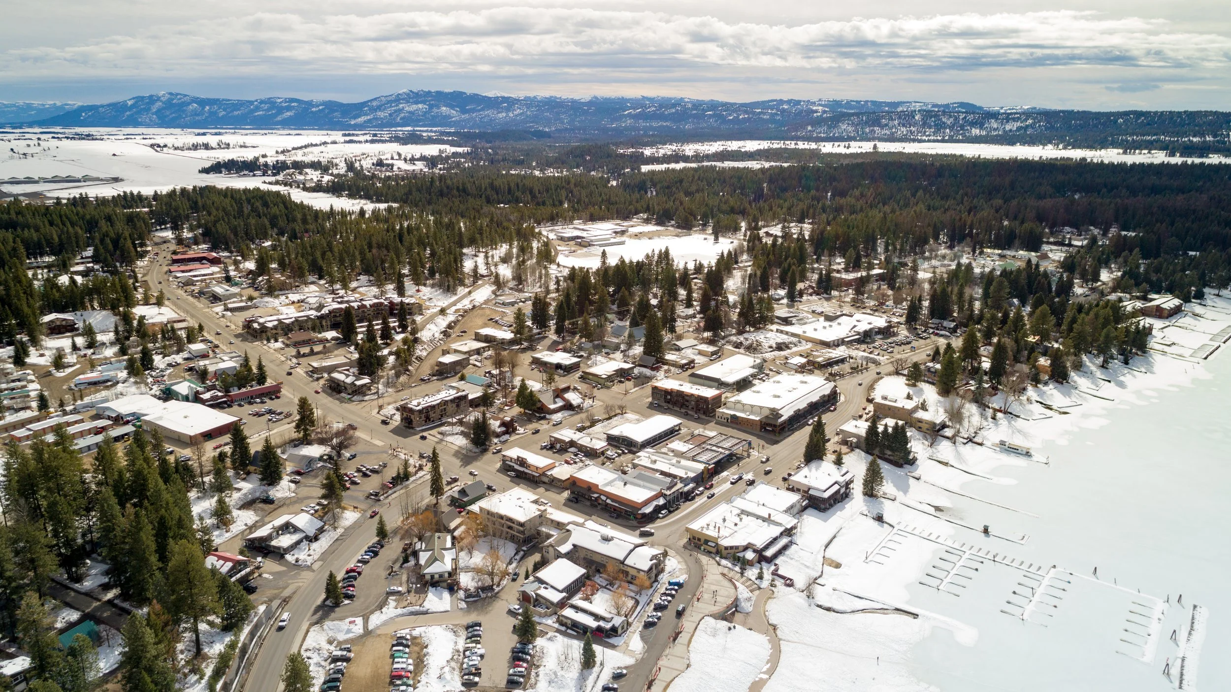Aerial view of a snow-covered downtown McCall near a frozen Lake Payette, with streets, trees, charming buildings, and mountains in the background.