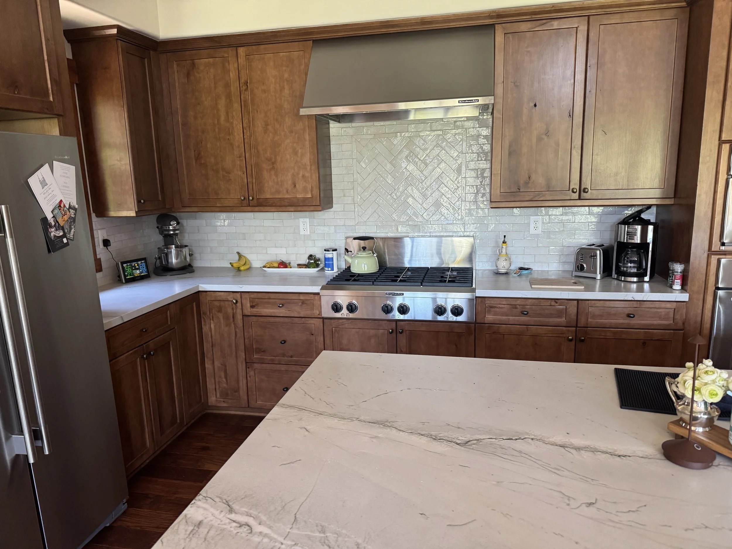 A kitchen with wooden cabinets, stainless steel oven and range, white marble countertops, a microwave, coffee maker, and various small items on the counter, including bananas and a clock.