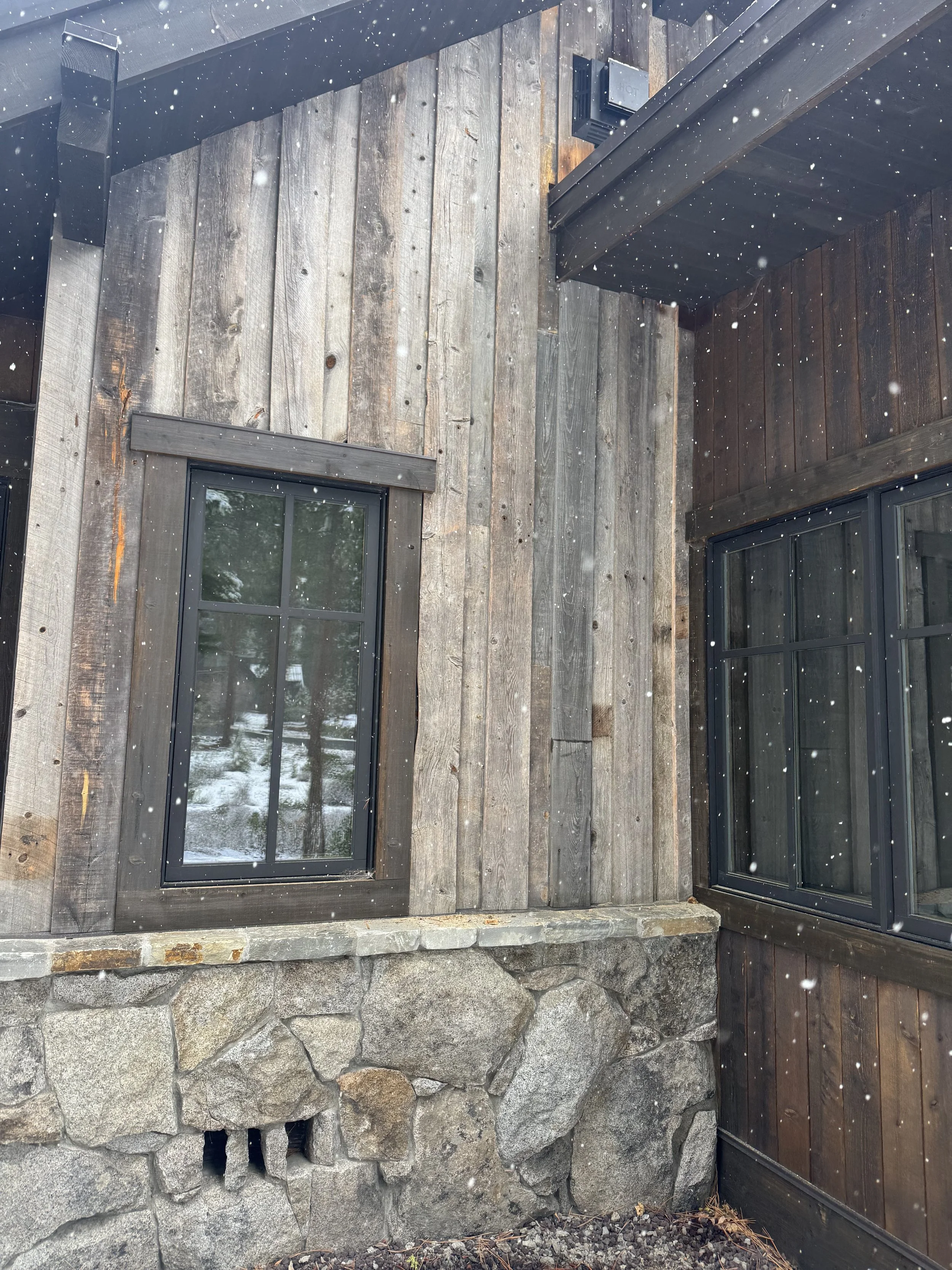 Close-up of a building exterior with wooden panel siding, two black-framed windows, a stone foundation, and a vent in the upper corner. Snow is falling.