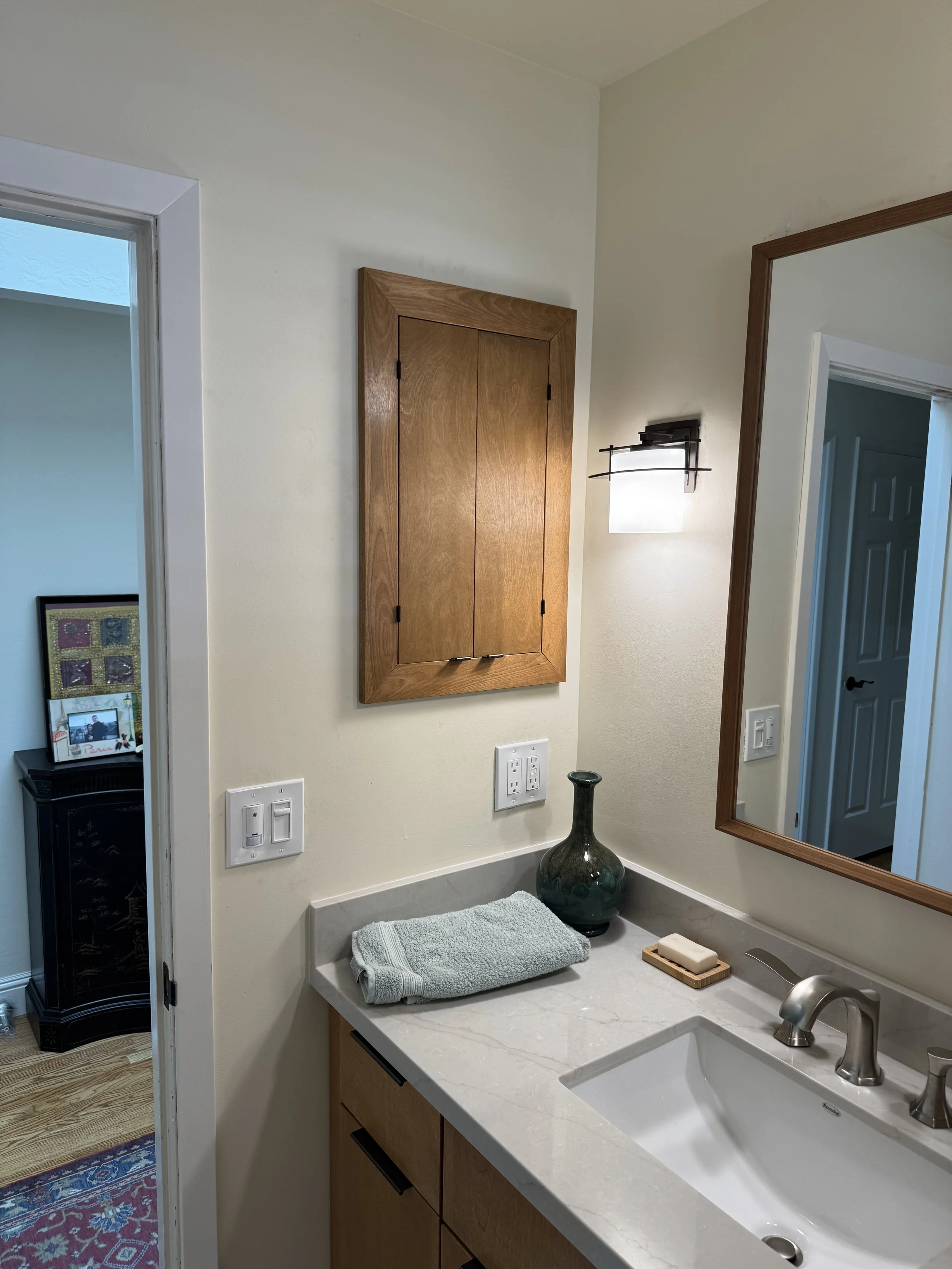 Bathroom corner with a white countertop, a soap dish, a bar of soap, a small vase, a mirror, a wall-mounted cabinet, eco-friendly towel, and light switches.