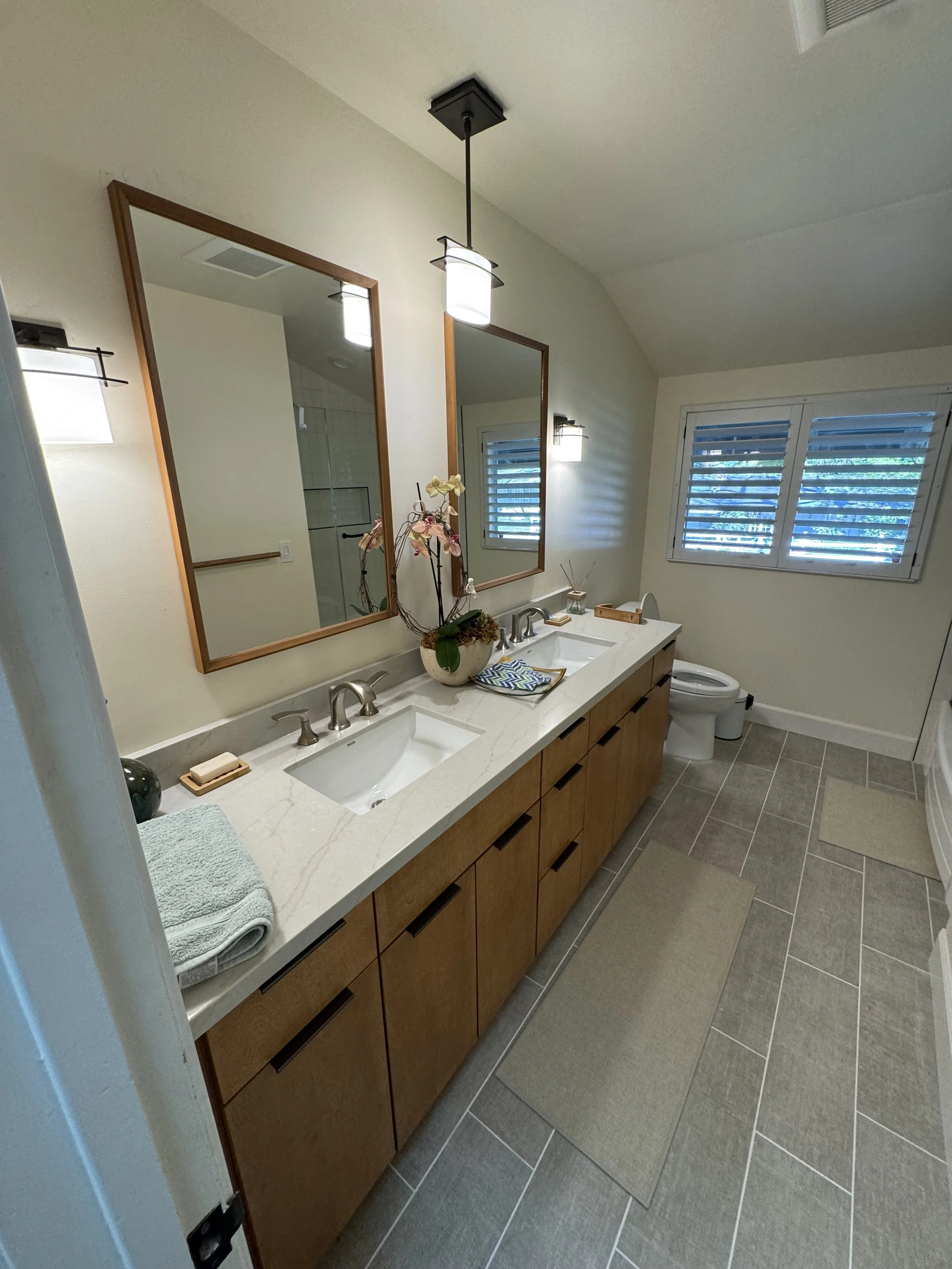 Double-sink bathroom vanity with wooden cabinets, decorated with a potted orchid, soap, and towels, mirror, window with blinds, and ceiling light fixture.