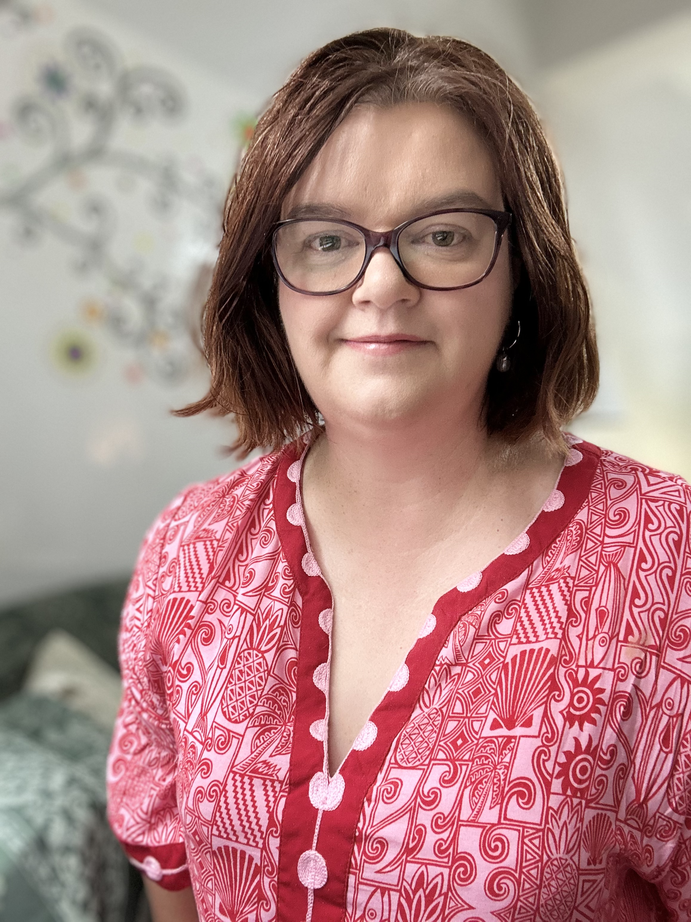A professional and approachable portrait of Leigh, Mental Health Nurse Practitioner with shoulder-length brown hair, glasses, and a pink patterned top standing in office space.
