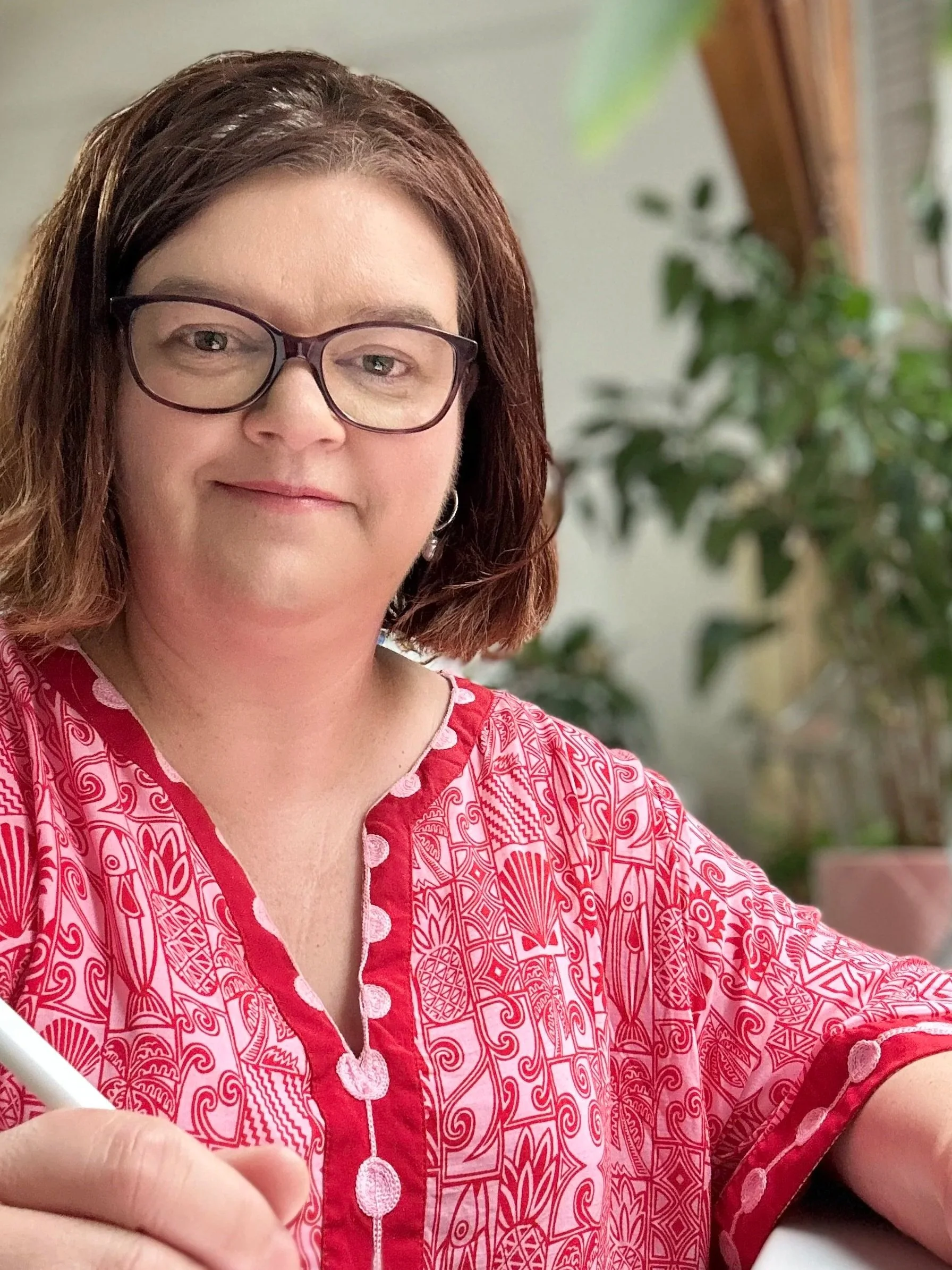 Your warm smiling female nurse practitioner sitting wearing glasses in a pink pattern dress, listening and scribbling.