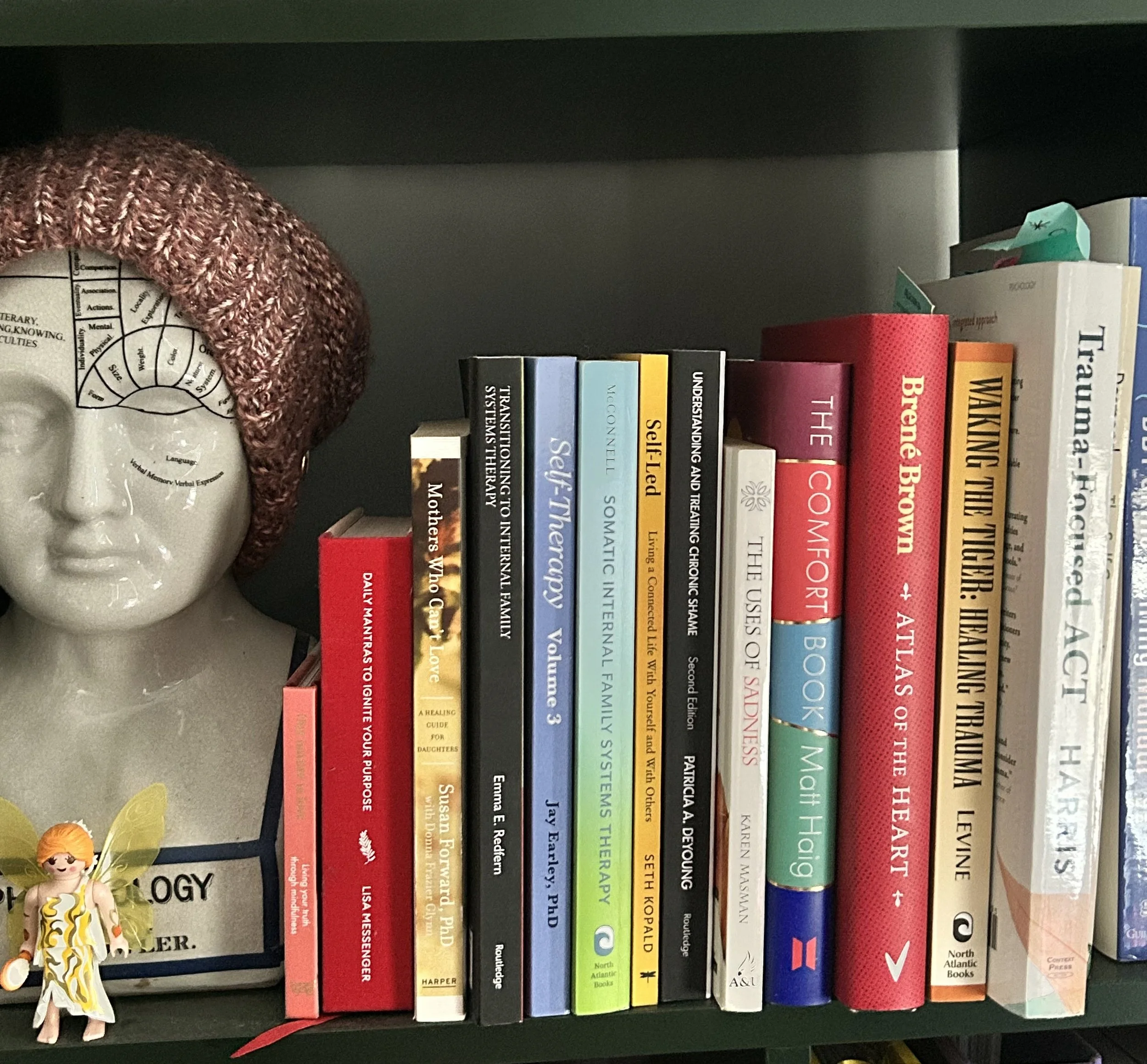 A bookshelf with a ceramic head sculpture wearing a brown knit hat, alongside various books on therapy.