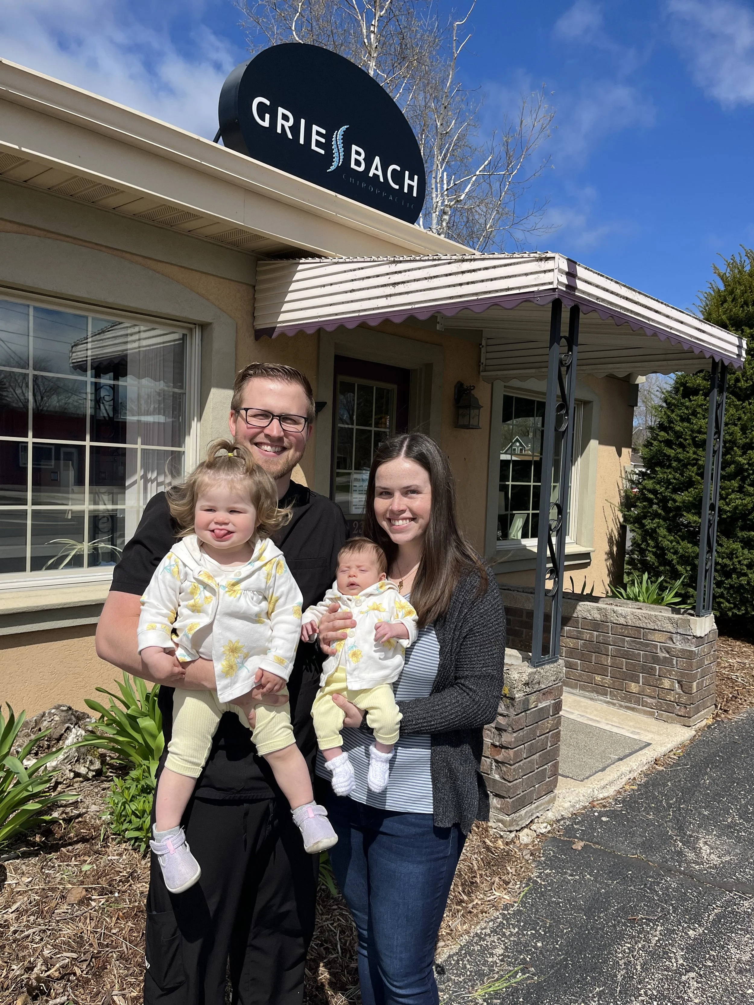 A smiling family of four standing outside a chiropractic office, with two young children, in front of the building with a sign that reads 'Grie Bach Chiropractic'.