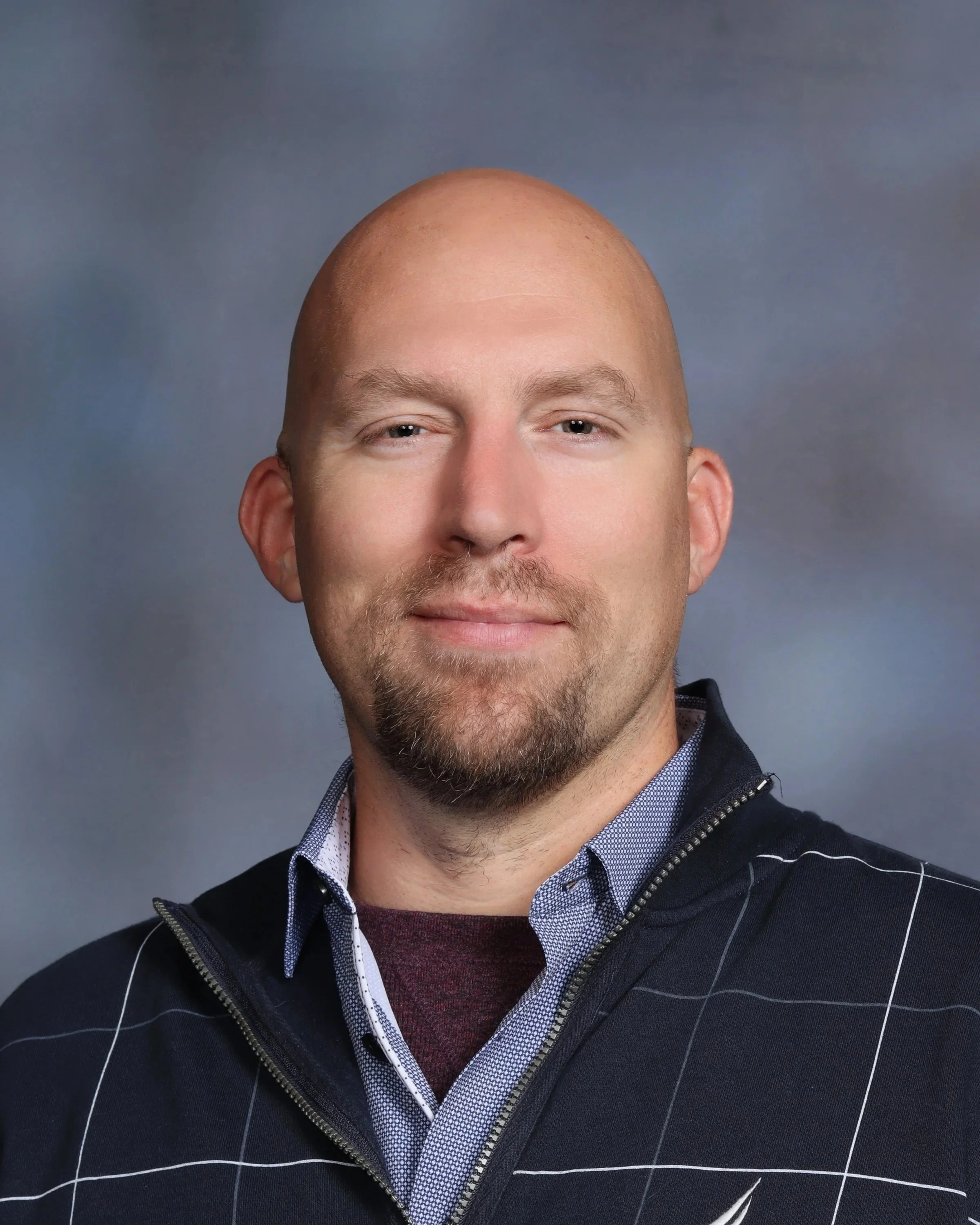 Headshot of a smiling bald man with light skin, wearing a dark jacket with white grid lines over a collared shirt, against a blurred gray background.