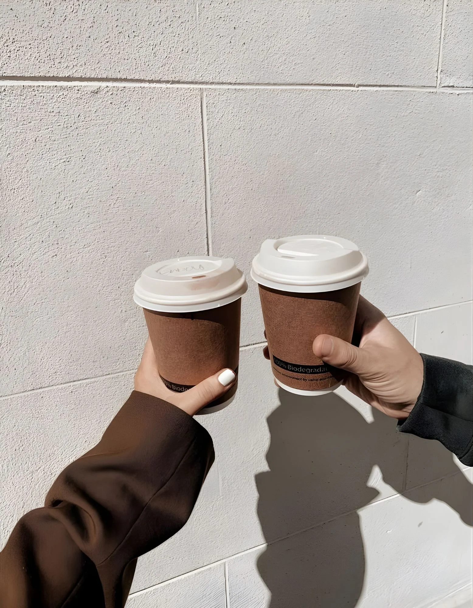 Two hands holding paper coffee cups in front of a light-colored tiled wall, representing love, connection and holistic wellbeing.