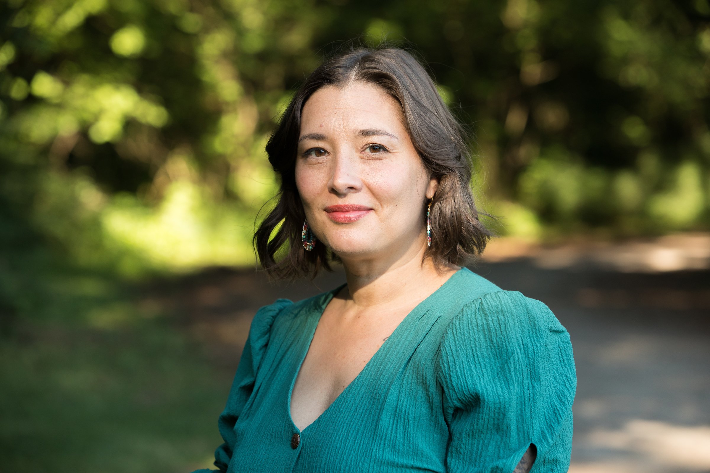 A woman with dark, wavy hair wearing a teal blouse and colorful earrings outdoors with a blurred green and brown forest background.