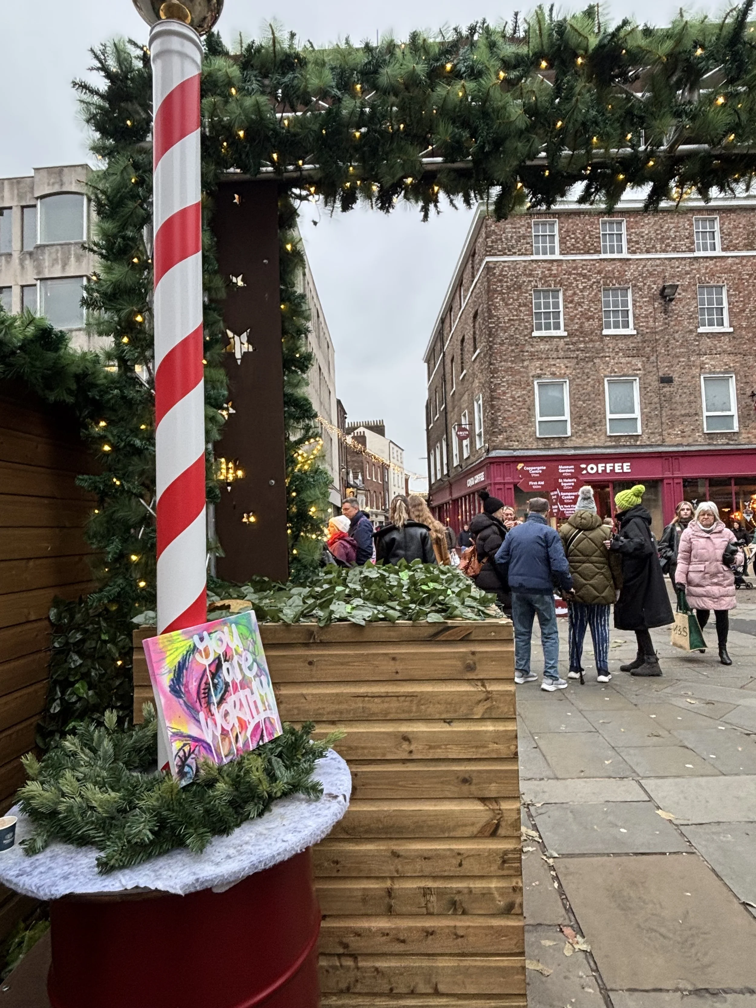 Christmas market scene on a city street with a wooden stall decorated with green garland and lights. There is a painted sign on the stall and a red and white striped pole. People dressed in winter clothing are gathered around, and brick and concrete 