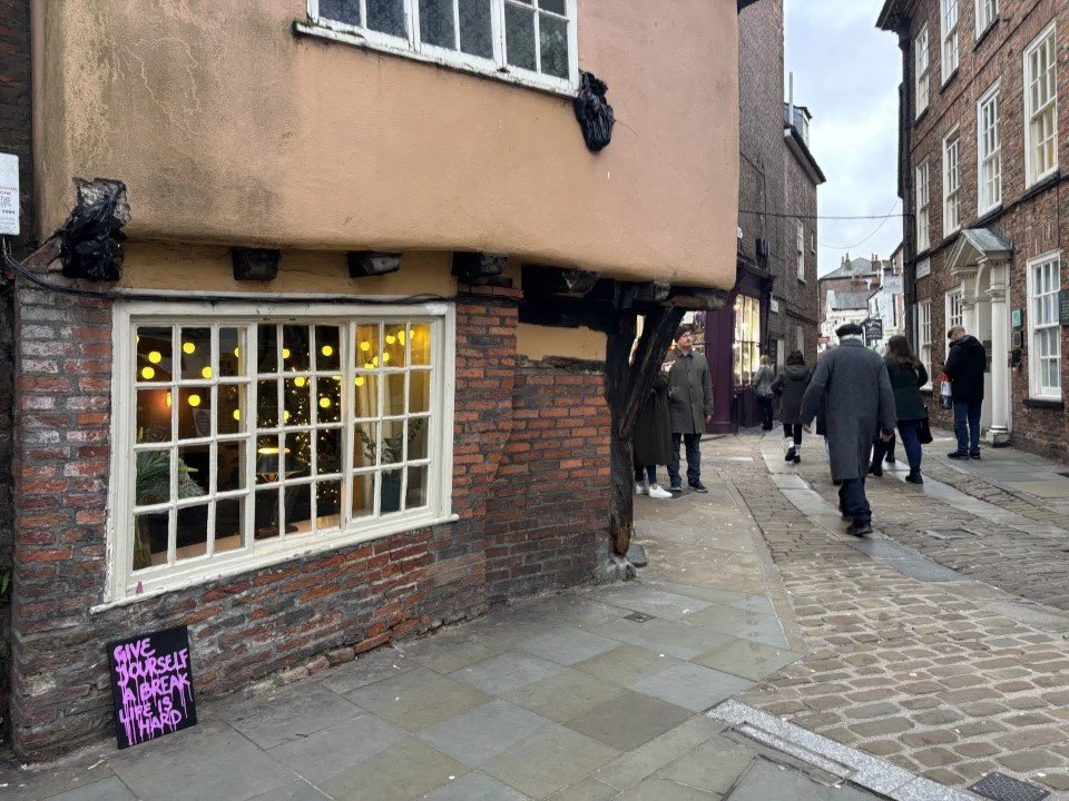 Street view with pedestrians walking past a building with a Christmas tree visible through a large front window. The window has a white grid frame, and the building has a mix of brick and plaster exterior. There is a small purple sign on the sidewalk
