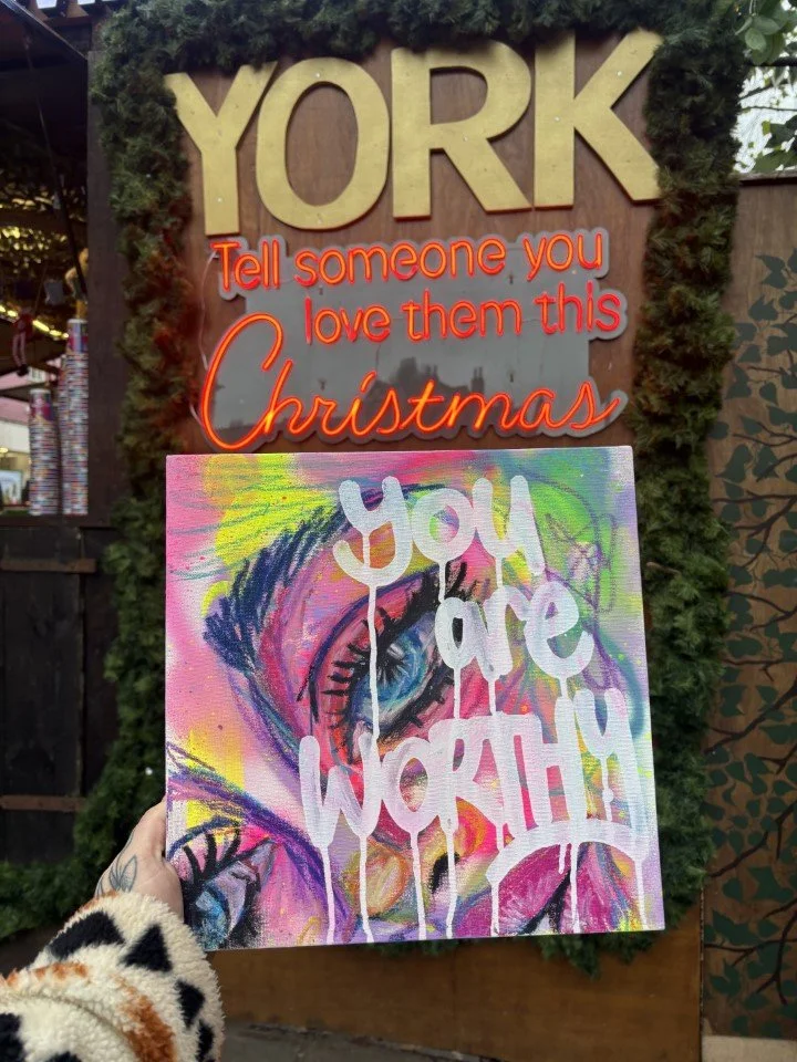 A person holding a colorful painting with the words 'You are WORTH' written across it, in front of a Christmas-themed display that says 'YORK' and 'Tell someone you love them this Christmas.'