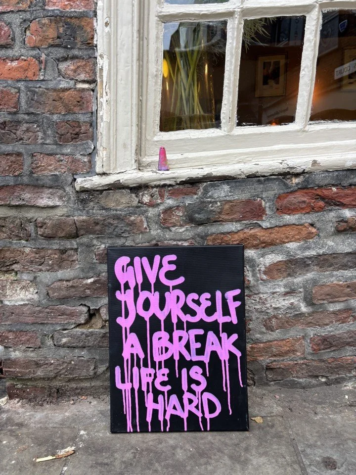 A black signboard with purple graffiti-style text that reads, "Give yourself a break. Life is hard." leaned against a brick wall outside a building, with a window above showing indoor plants and framed pictures.