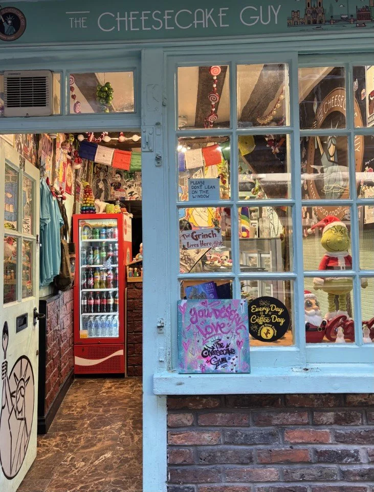 Exterior view of a bakery or café named 'The Cheesecake Guy' with a blue entrance, decorated inside with colorful ornaments, signs, and a Christmas-themed display, including a stuffed elf.
