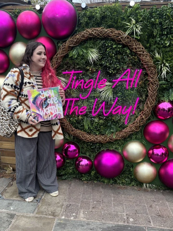 Woman with pink hair in a patterned sweater, striped pants, and white shoes holding a colorful art piece, standing next to a holiday-themed decorative wall with large pink and gold ornaments, green foliage, and a neon sign that says "Jingle All The W