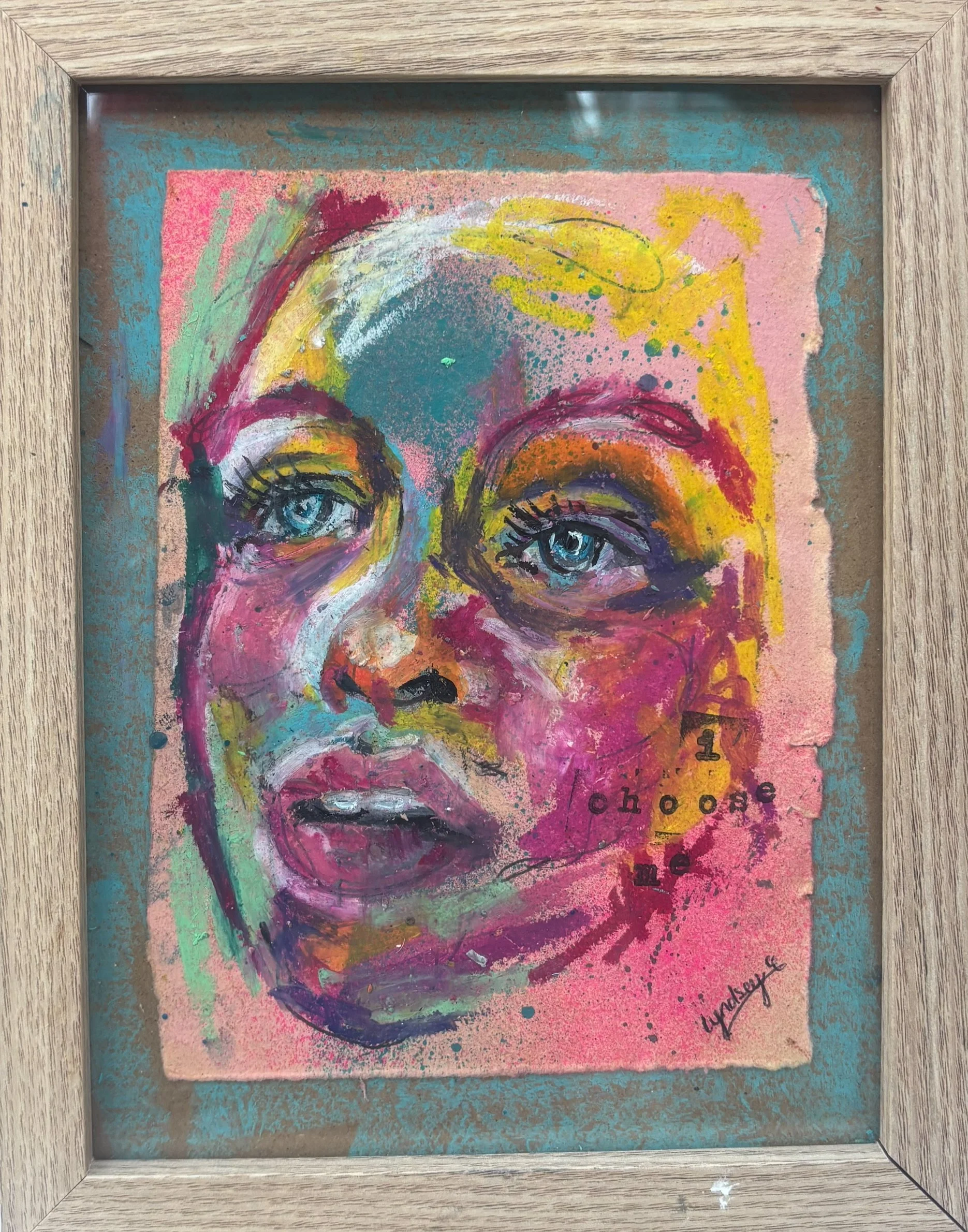 A colorful abstract portrait of a woman's face with prominent blue eyes, pink cheeks, and lips, surrounded by splashes of vibrant colors like yellow, pink, green, and purple, framed in wood.