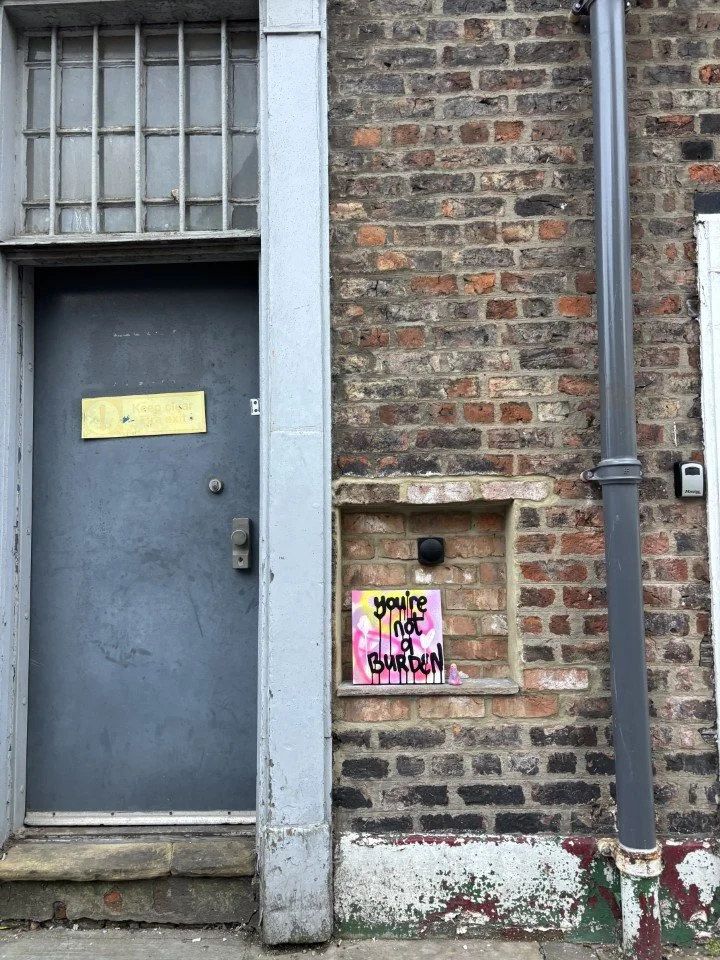 Brick wall with a gray door on the left, a metal window above, and a gray drainpipe on the right. There is graffiti artwork that says 'You're not a burden' on a small ledge in the brick wall.