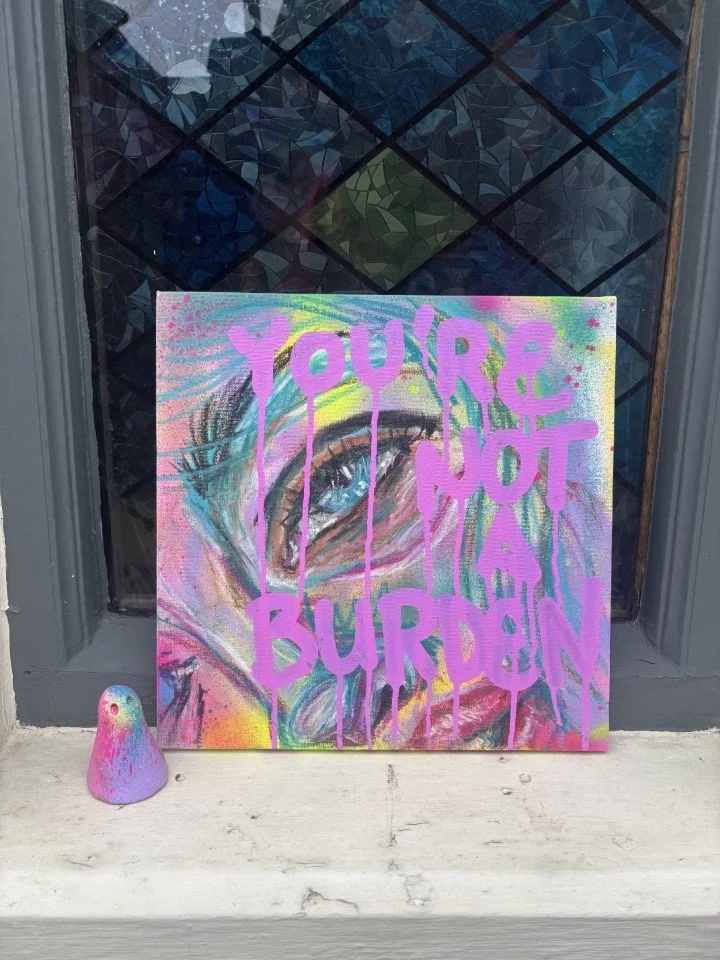 A colorful graffiti-style painting of a human eye with the words "You're not your burden" written in purple paint over the image, placed on a sidewalk in front of a black textured window.