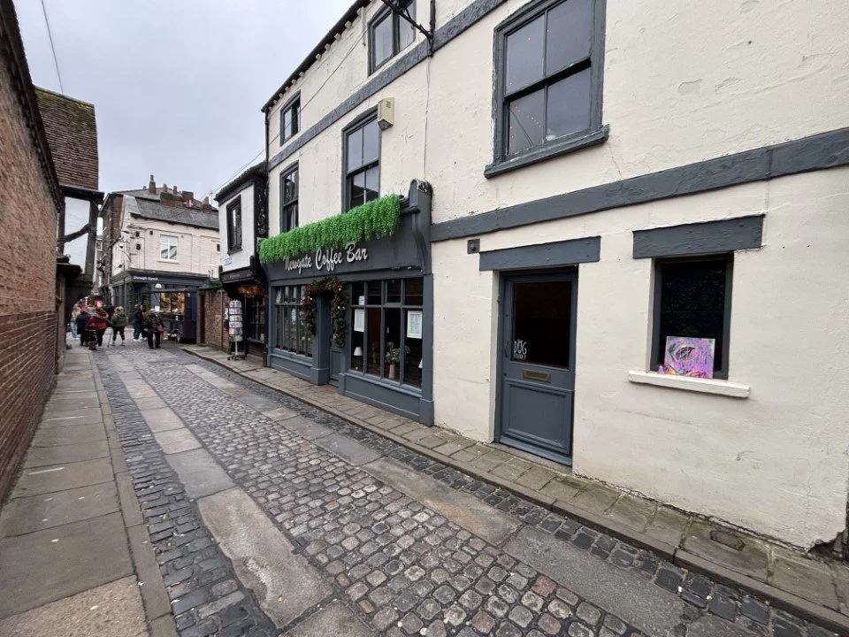 A narrow cobblestone street lined with buildings, including a cafe with a green sign and an outdoor seating area, and a group of people walking further down the street on an overcast day.