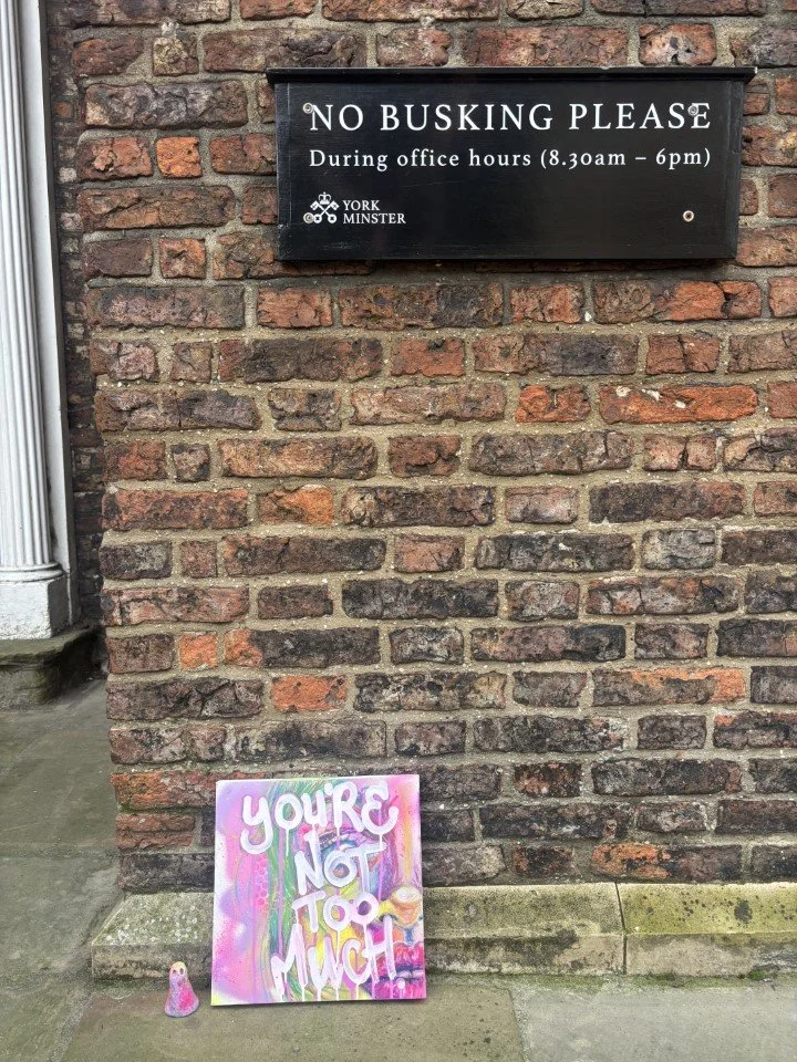A black sign on a brick wall reads 'NO BUSKING PLEASE During office hours (8:30am - 6pm)' with the York Minster logo. Below, on the sidewalk, is a colorful sign with graffiti-style text that says 'YOU'RE NOT TOO MUCH' and a small pink object next to 