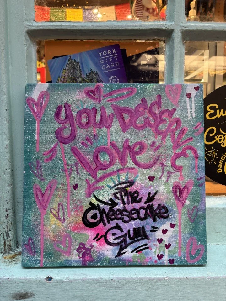 Colorful painting with pink and purple words saying "You Deserve Love" and "The Chosecape Guy" surrounded by pink hearts and squiggles, across a pastel background.