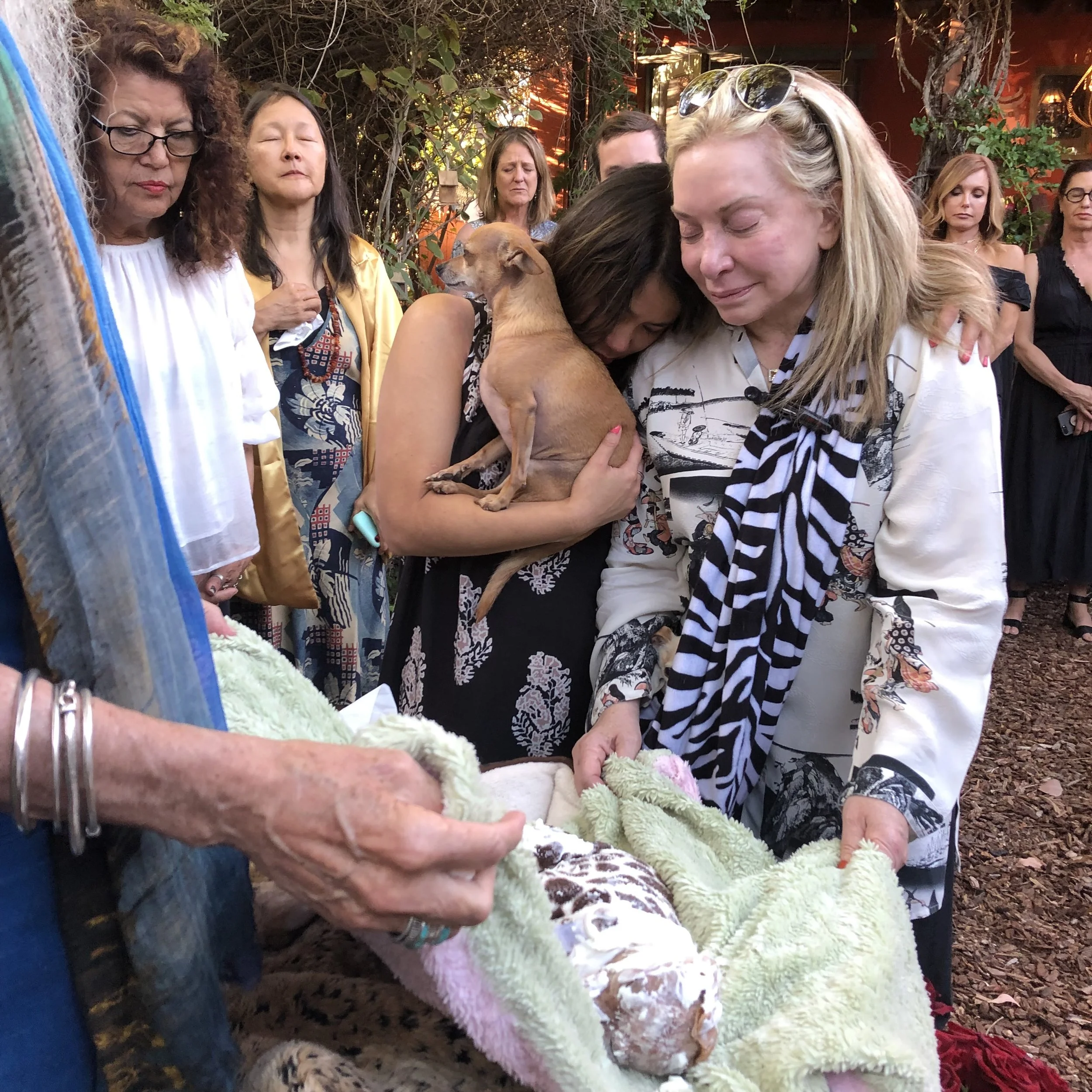 A group of people gathered outdoors for a pet funeral, with two women in the foreground holding a deceased dog in a blanket. One woman is holding a small dog close to her chest. The group appears emotional during a special moment at a pet funeral.