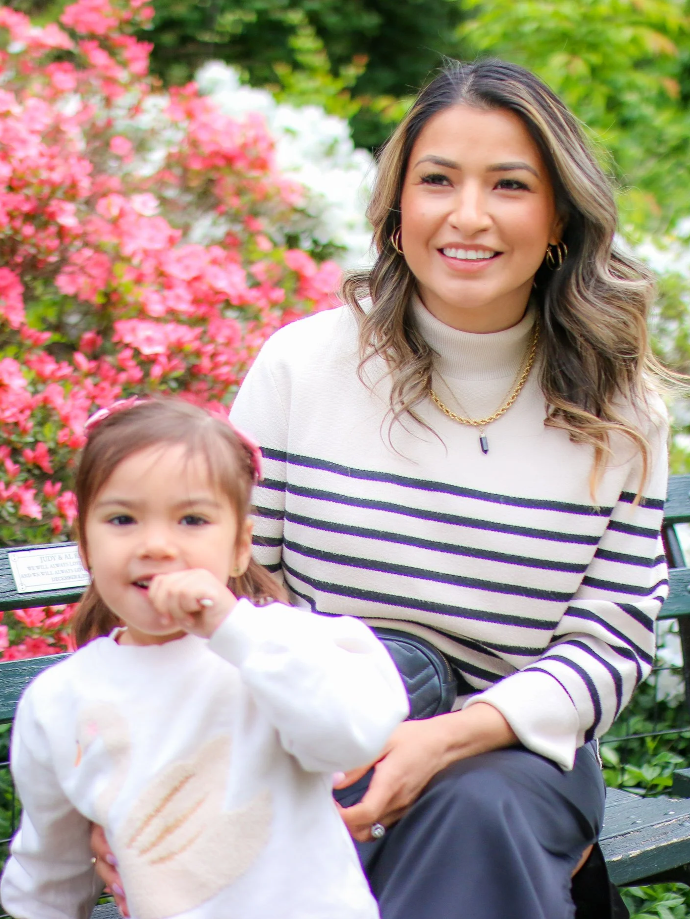 A woman sitting on a park bench with a young girl, greenery and pink flowers in the background. The woman has shoulder-length wavy hair, wearing a striped sweater, gold jewelry, and has a smile. The girl has brown pigtails, is wearing a white sweater, and is pointing towards the camera.