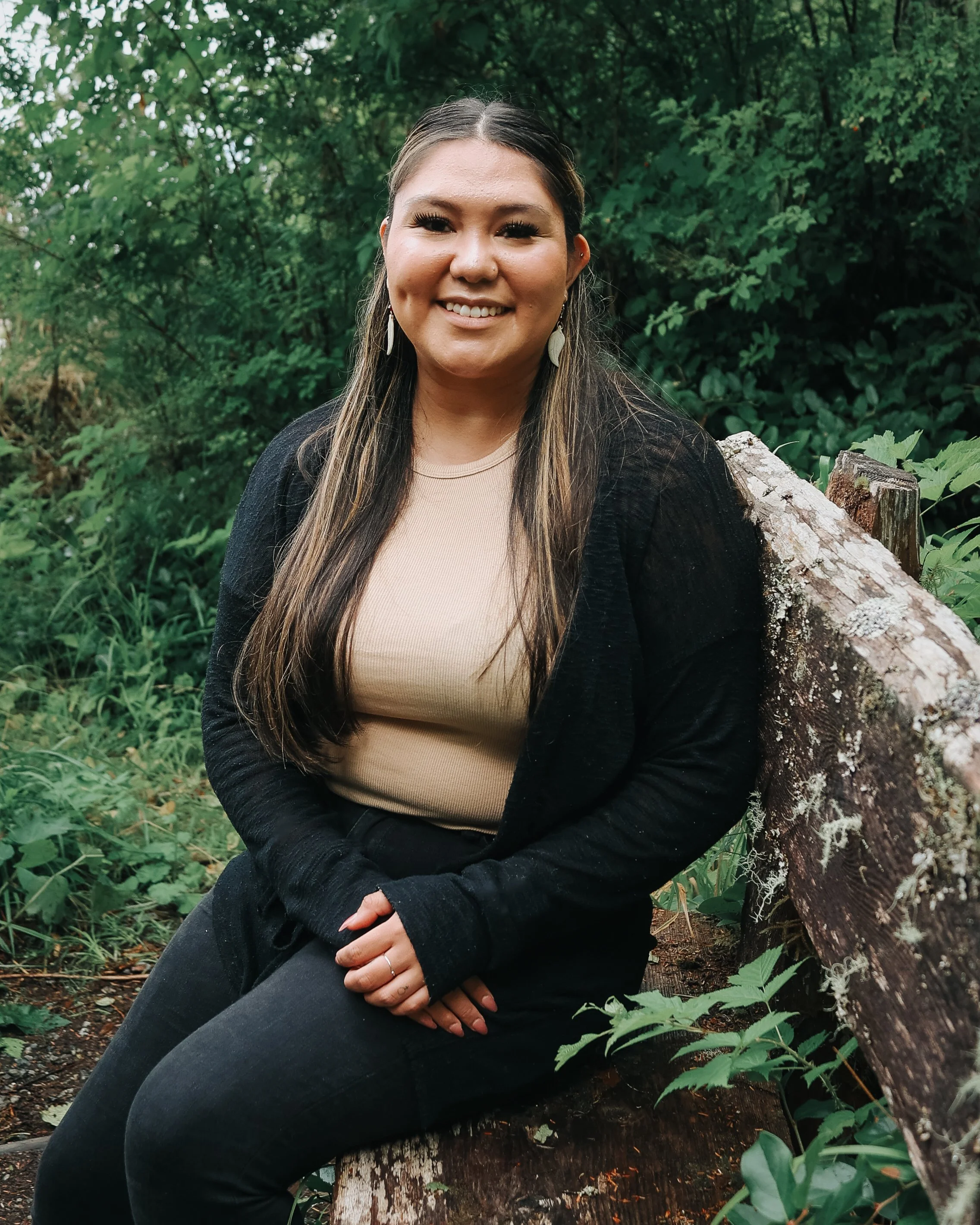 A smiling woman sitting on a fallen tree trunk in a green forest, wearing a beige top, black jacket, black pants, and earrings.