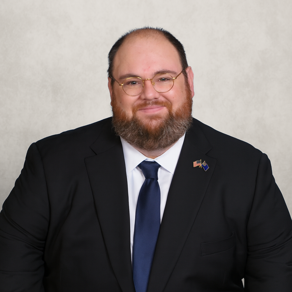 Professional portrait of Michael MacDougall, candidate for Nevada Controller, with glasses and a beard, wearing a suit and tie, with patriotic pins on his lapel, against a neutral background.