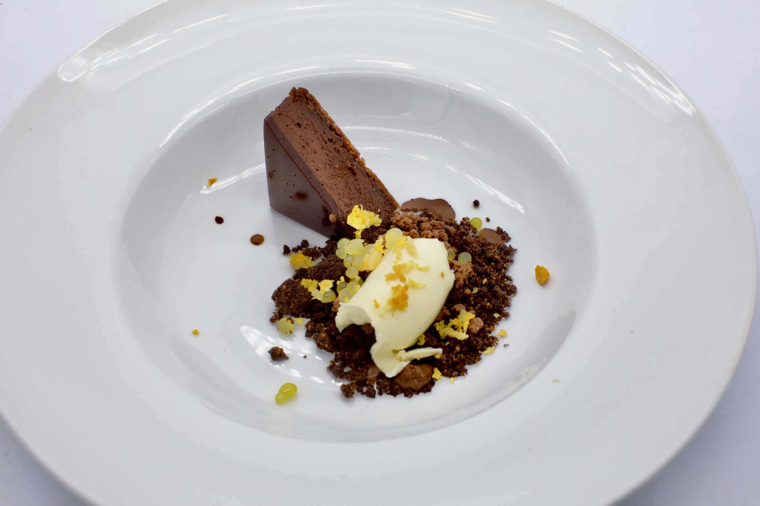 Dessert with a chocolate cake wedge, white cream, yellow crumbs, and chocolate sauce on a white plate.