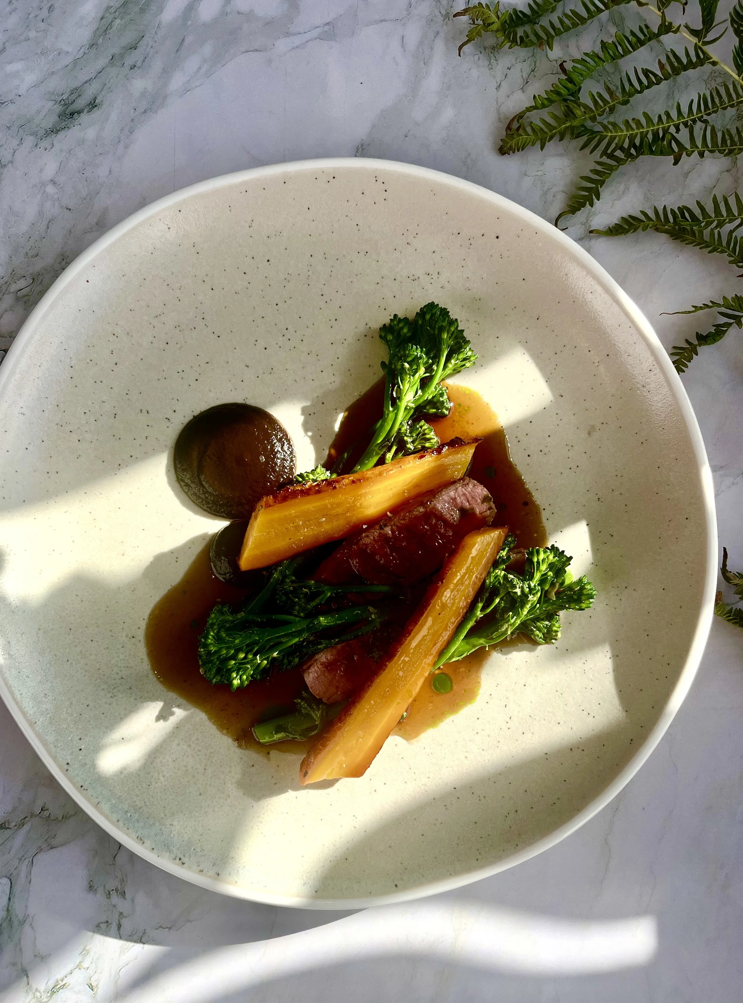 A white bowl with cooked broccoli, yellow vegetable sticks, a round dark brown item, and a sauce on a marble surface with fern leaves nearby.