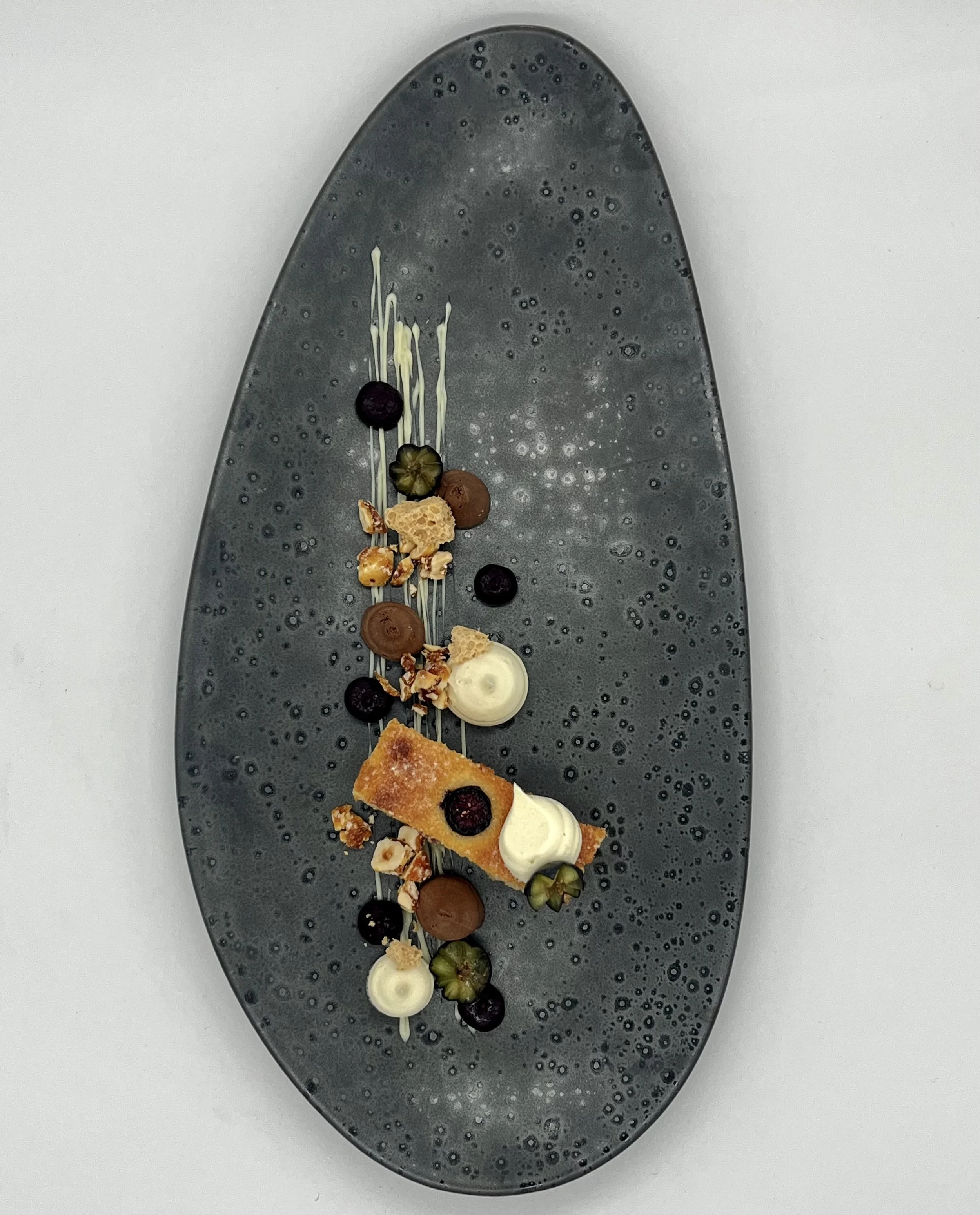 A dessert plate with a rectangular caramelized pastry, blueberries, small dollops of white cream, chopped nuts, and chocolate spheres on a dark, textured oval plate.