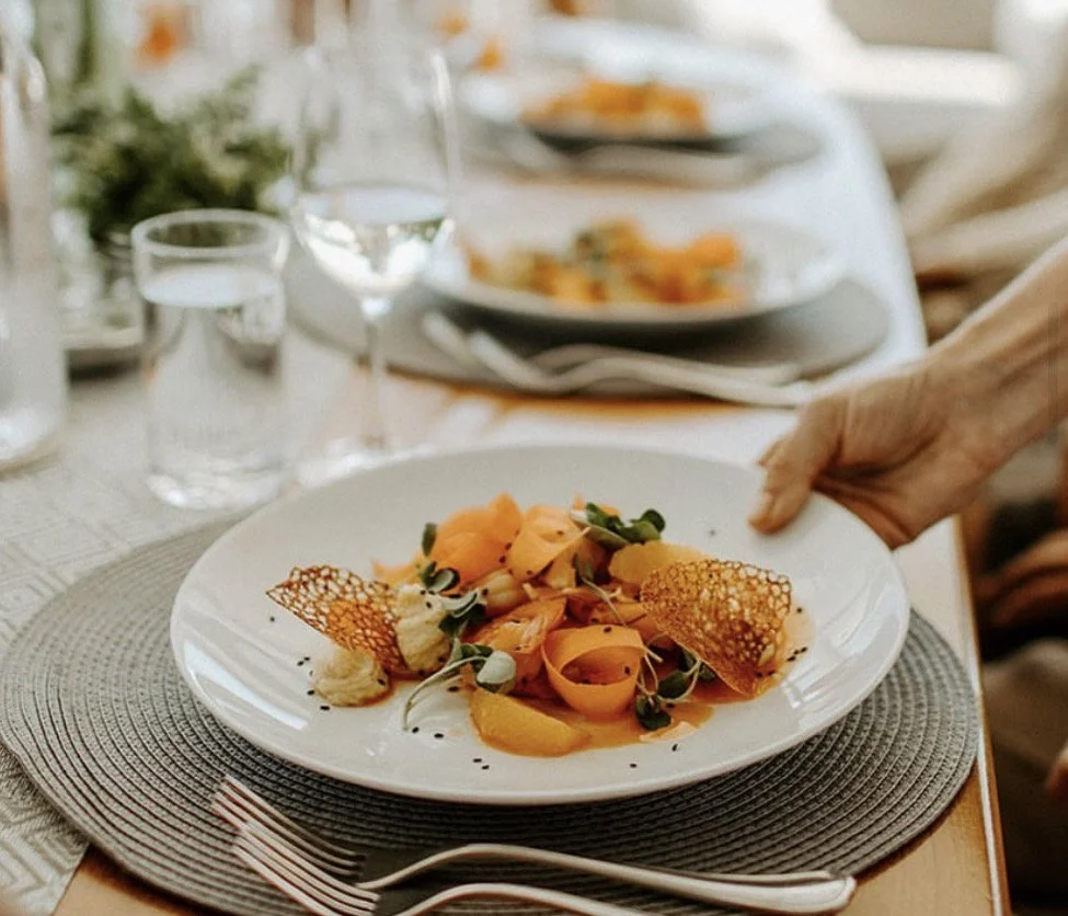 A person is serving a plated dish of thinly sliced vegetables and garnishes on a white plate at a formal dining table.