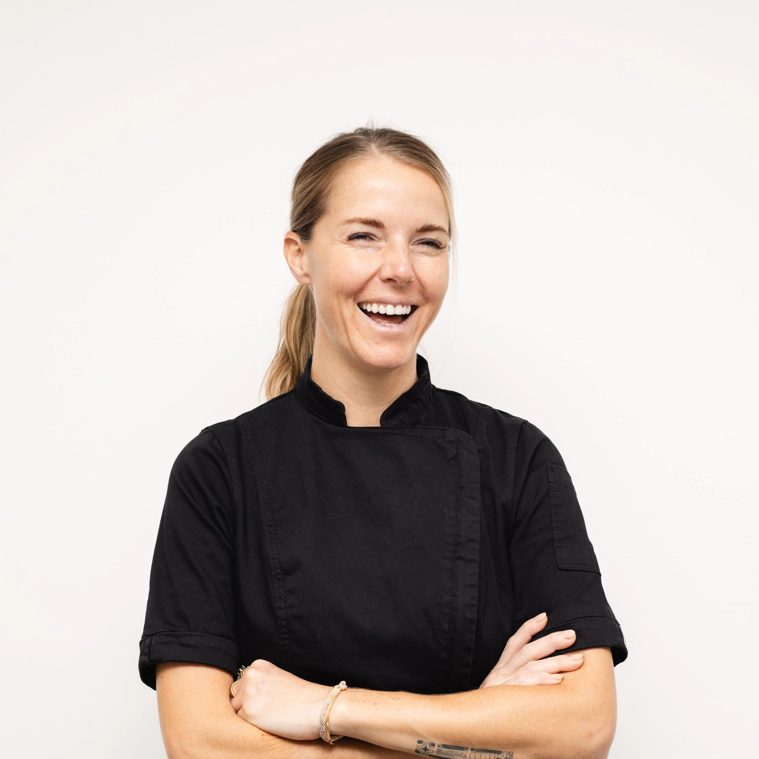 A woman with blonde hair in a ponytail, smiling with crossed arms, wearing a black chef's coat, standing against a plain white background.