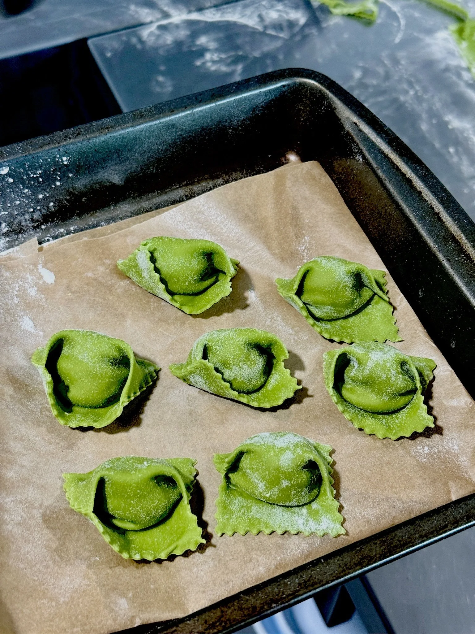 Uncooked green pasta filled with a filling, arranged on parchment paper on a baking tray, dusted with flour.