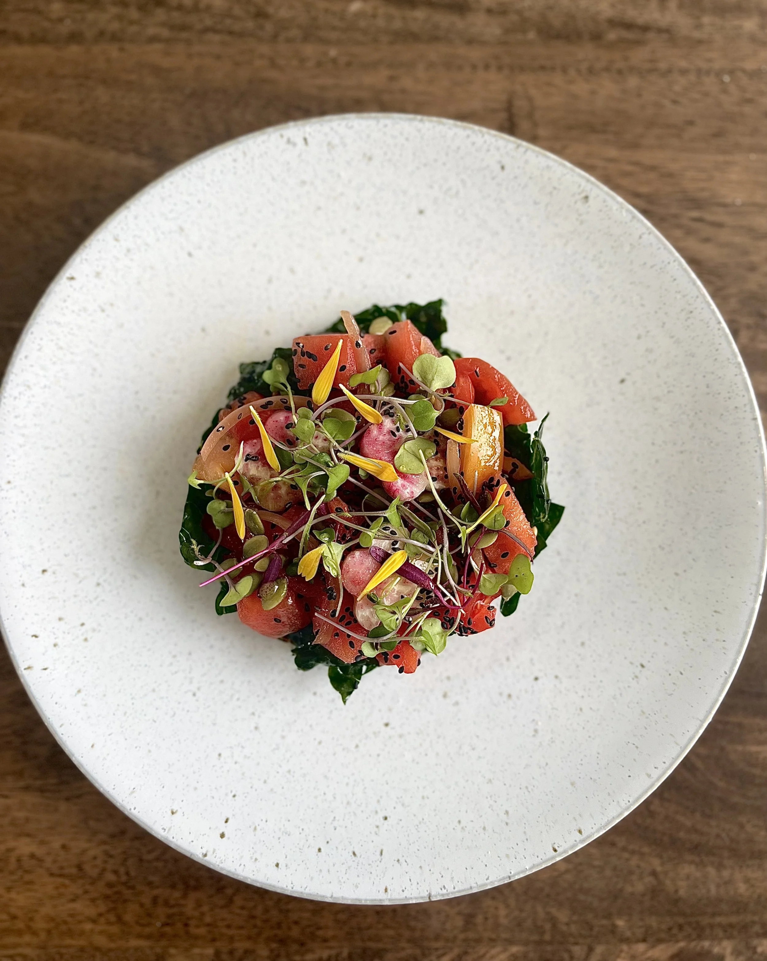A top-down view of a colorful salad with microgreens, chopped vegetables, and edible flowers on a white plate.