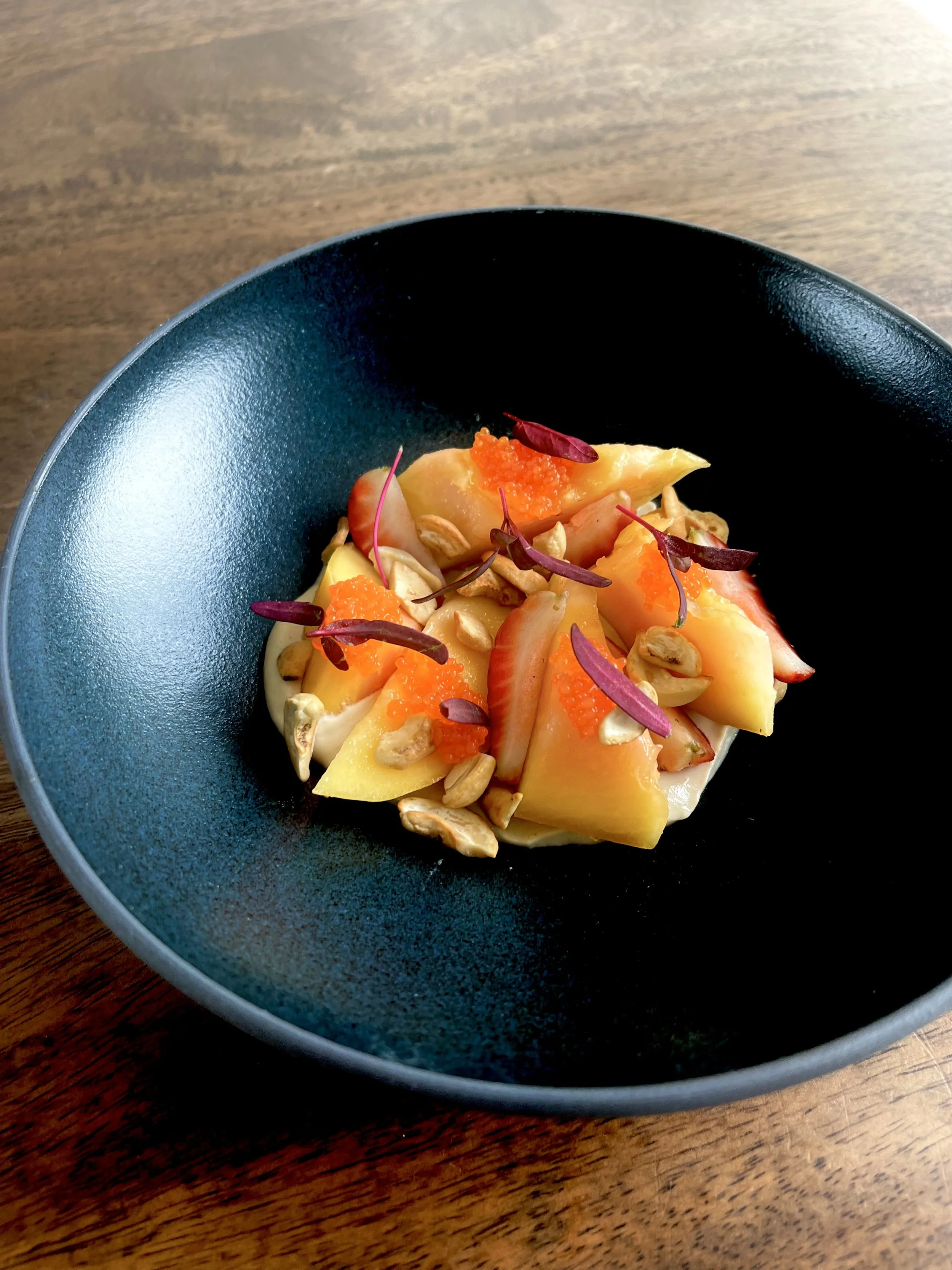A black bowl containing a fruit salad with sliced strawberries, peaches, and diced fruit, topped with sunflower seeds, orange fish roe, and purple microgreens, placed on a wooden surface.