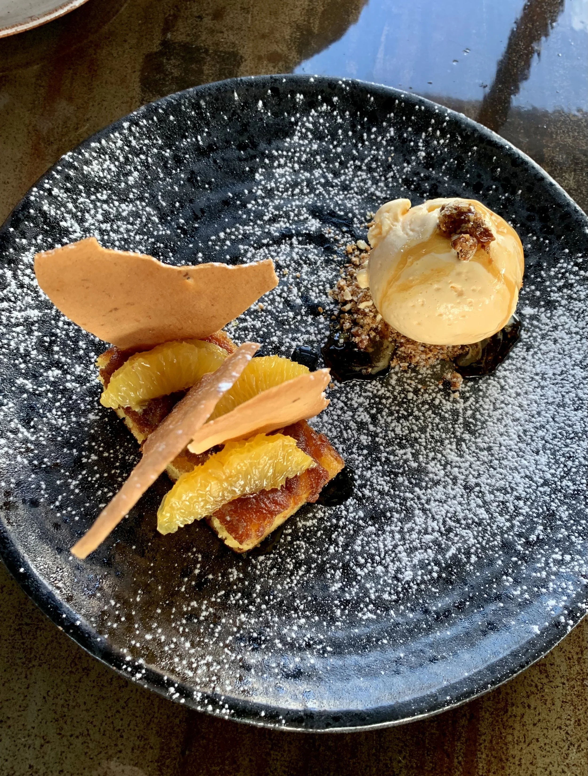 Dessert with orange slices, a crispy biscuit, and a scoop of ice cream topped with caramel and nuts on a black plate dusted with powdered sugar.