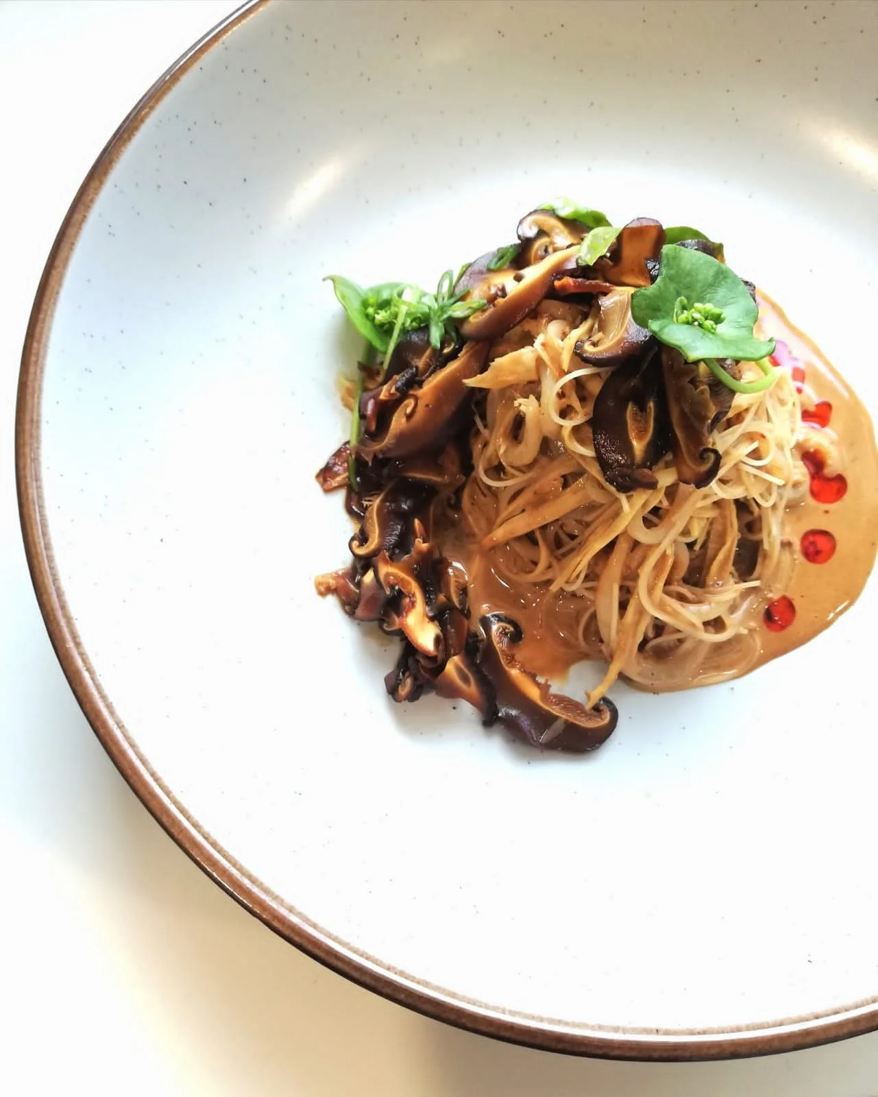 A plate of Asian-style noodles topped with sautéed shiitake mushrooms, garnished with microgreens and red pepper flakes.