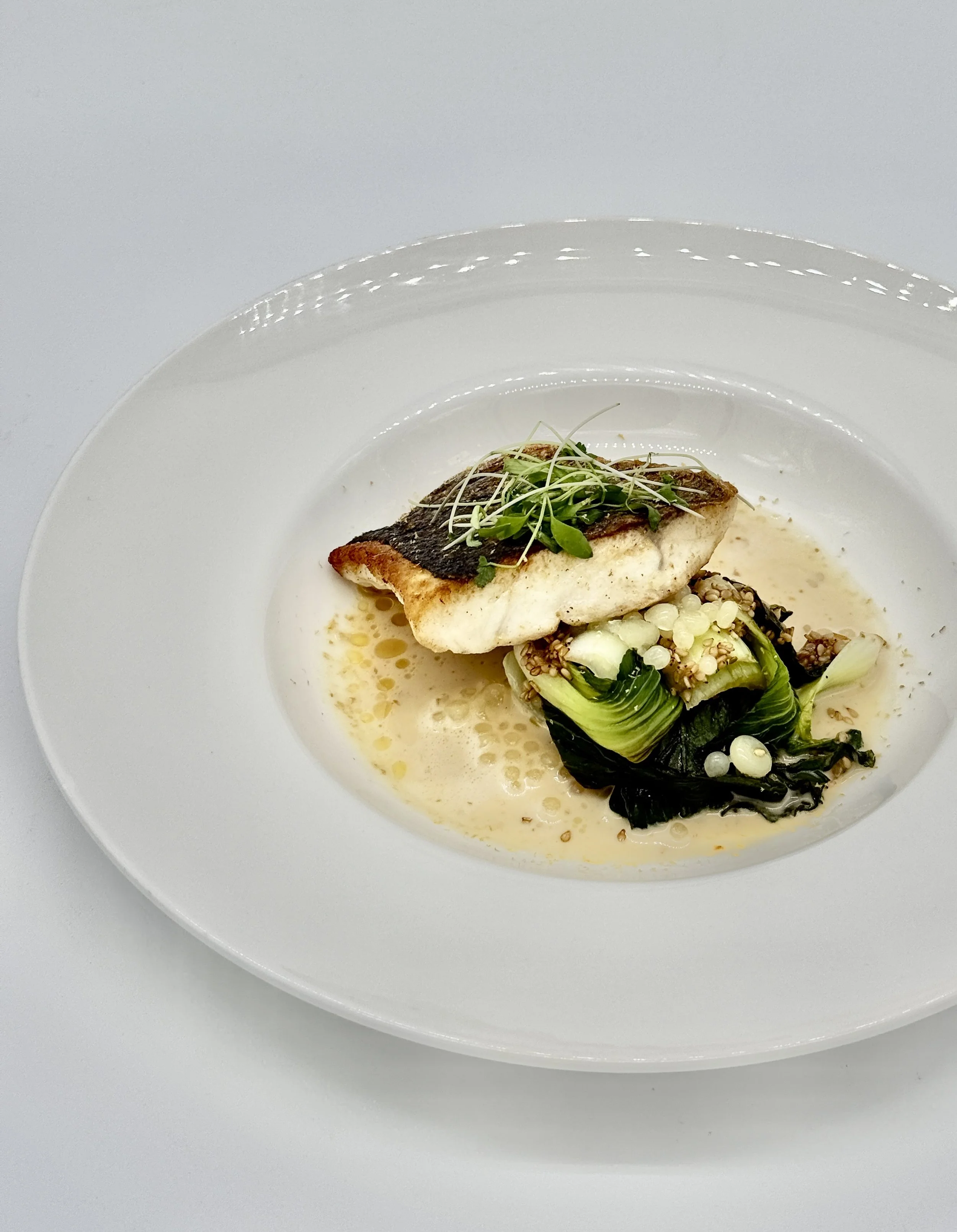 A plated dish with a piece of cooked fish garnished with microgreens and accompanied by cooked greens and grains, with a light sauce.