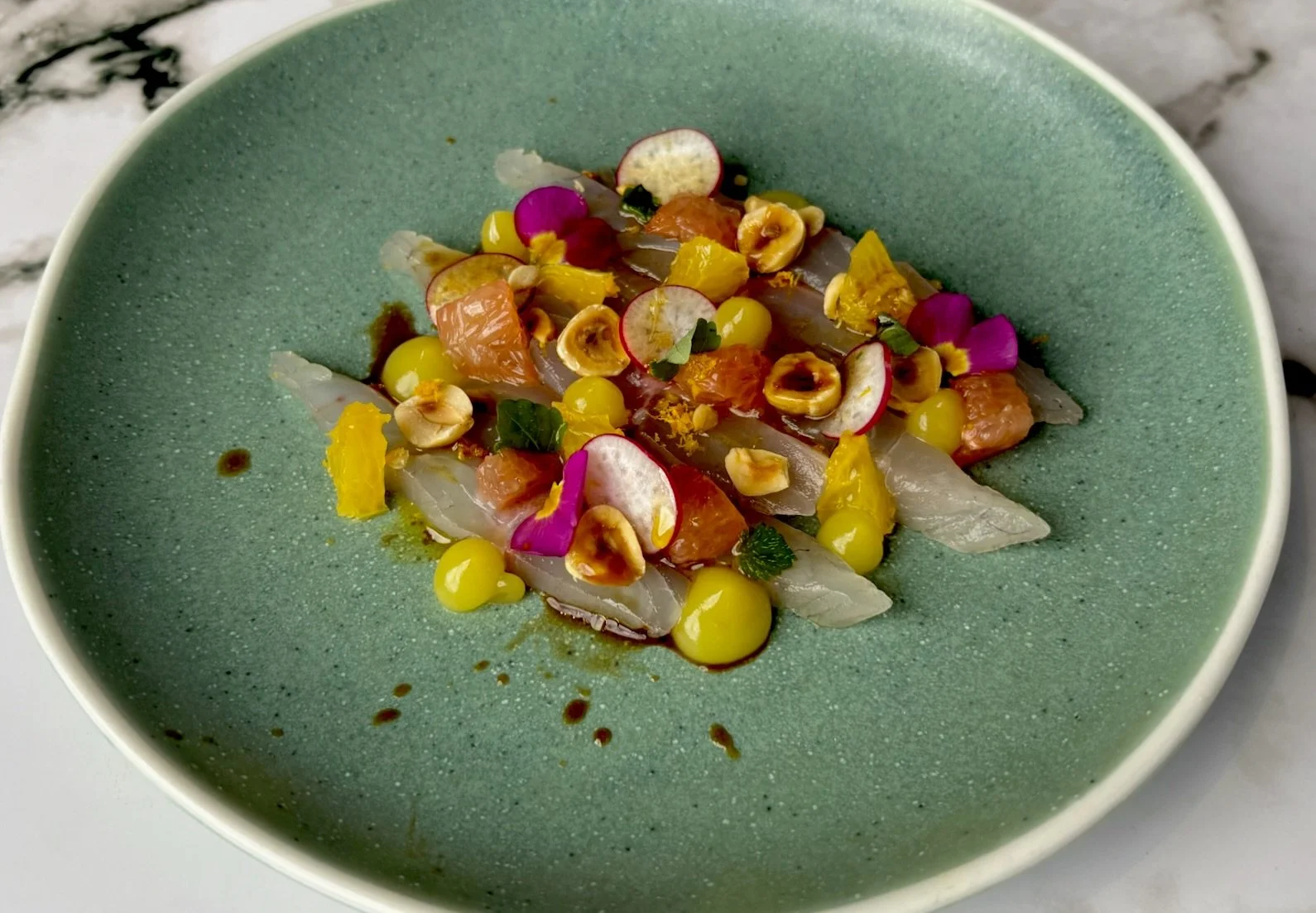 A colorful fish ceviche dish with sliced radishes, yellow berries, orange citrus, chopped herbs, and edible flower petals, served in a green bowl.