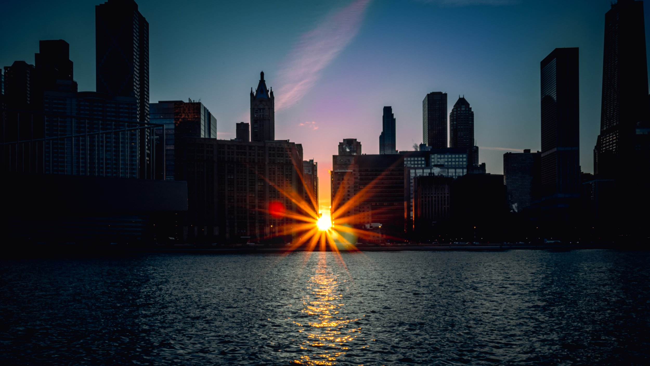 Sunset over a city skyline with tall skyscrapers, reflecting off the river in the foreground.