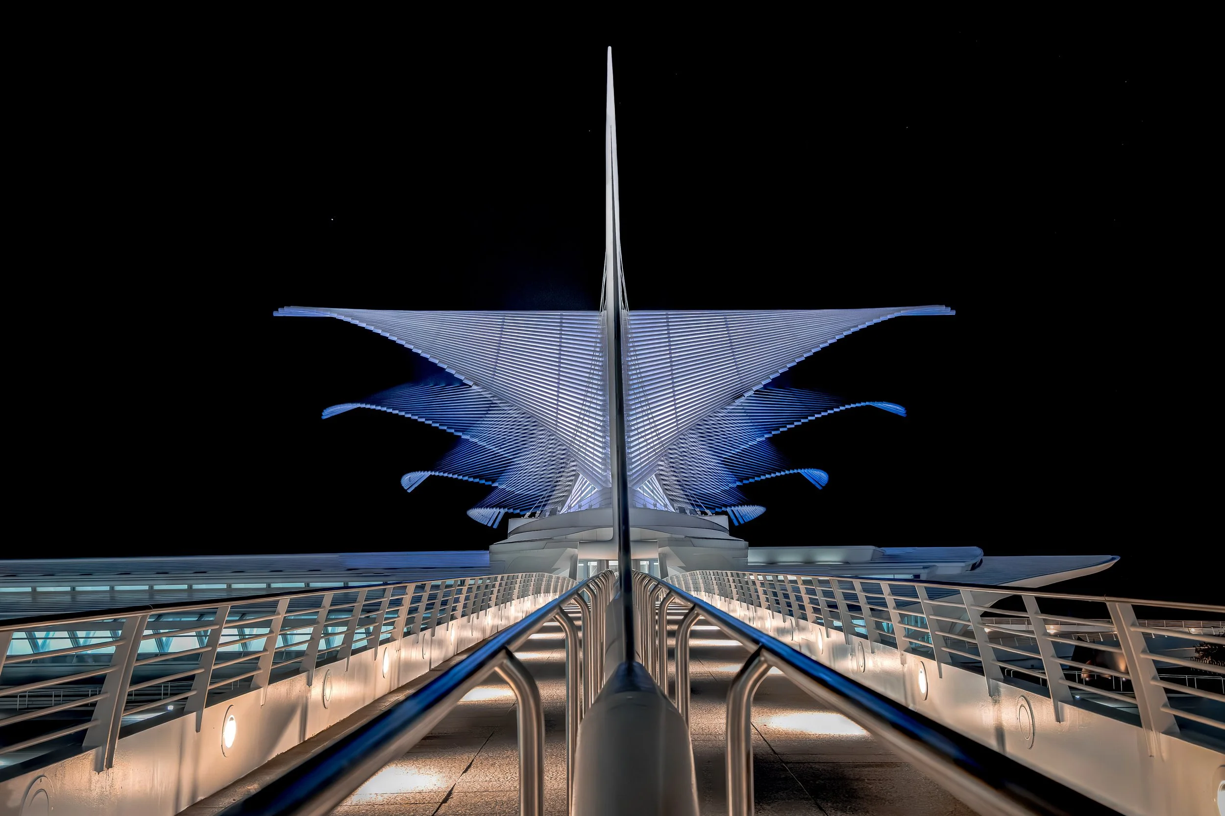Futuristic bridge at night with illuminated, wing-like structures extending outward, and a central sharp spire, captured from the walkway between railings.